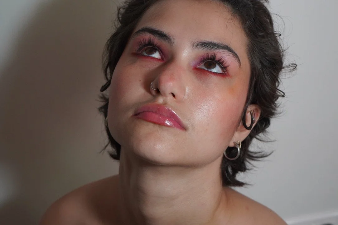 A person with short dark hair, wearing multiple earrings, looking upwards with eyes slightly rolled back, wearing shiny pink lipstick and eye makeup.