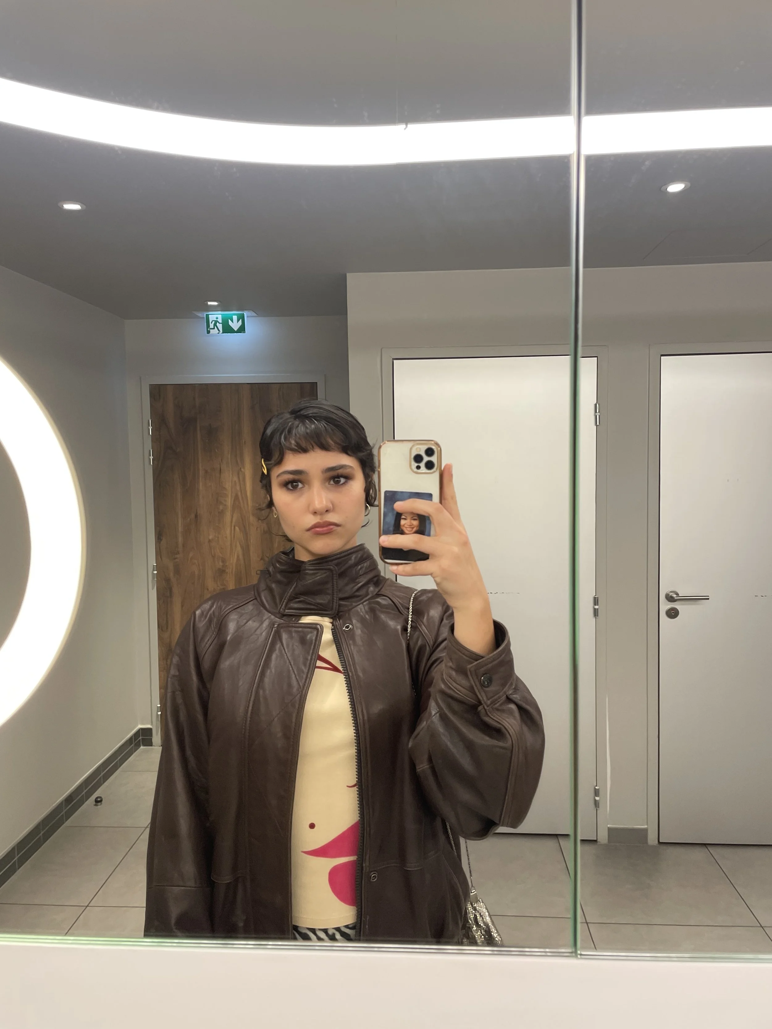 A young woman taking a mirror selfie in a bathroom or restroom, wearing a brown leather jacket over a cream-colored top with red and pink design, with a wooden door and white walls in the background.