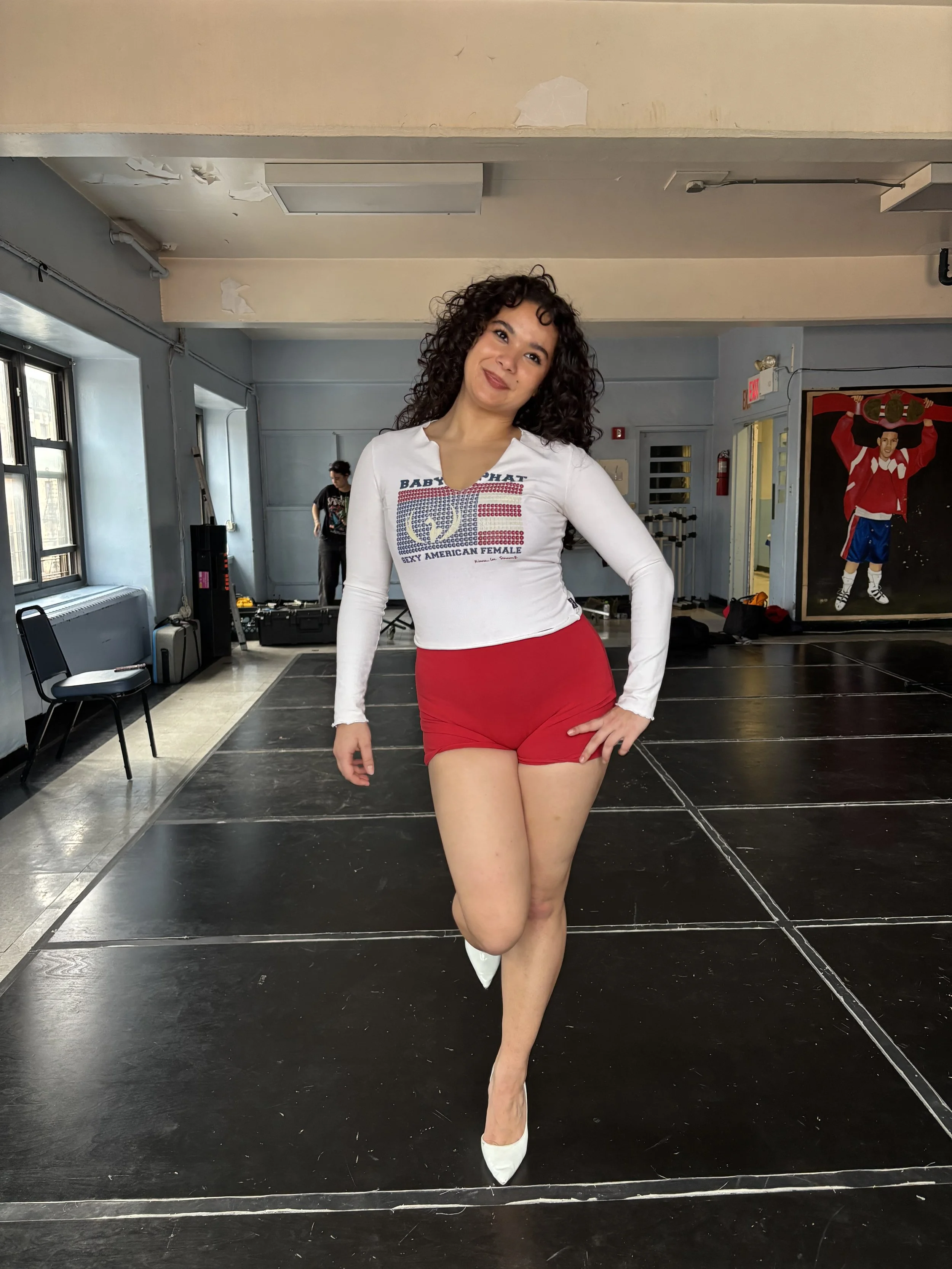 A woman with curly dark hair posing on a dance studio stage, wearing a white long-sleeve shirt with American flag and text design, red shorts, and white high heels.