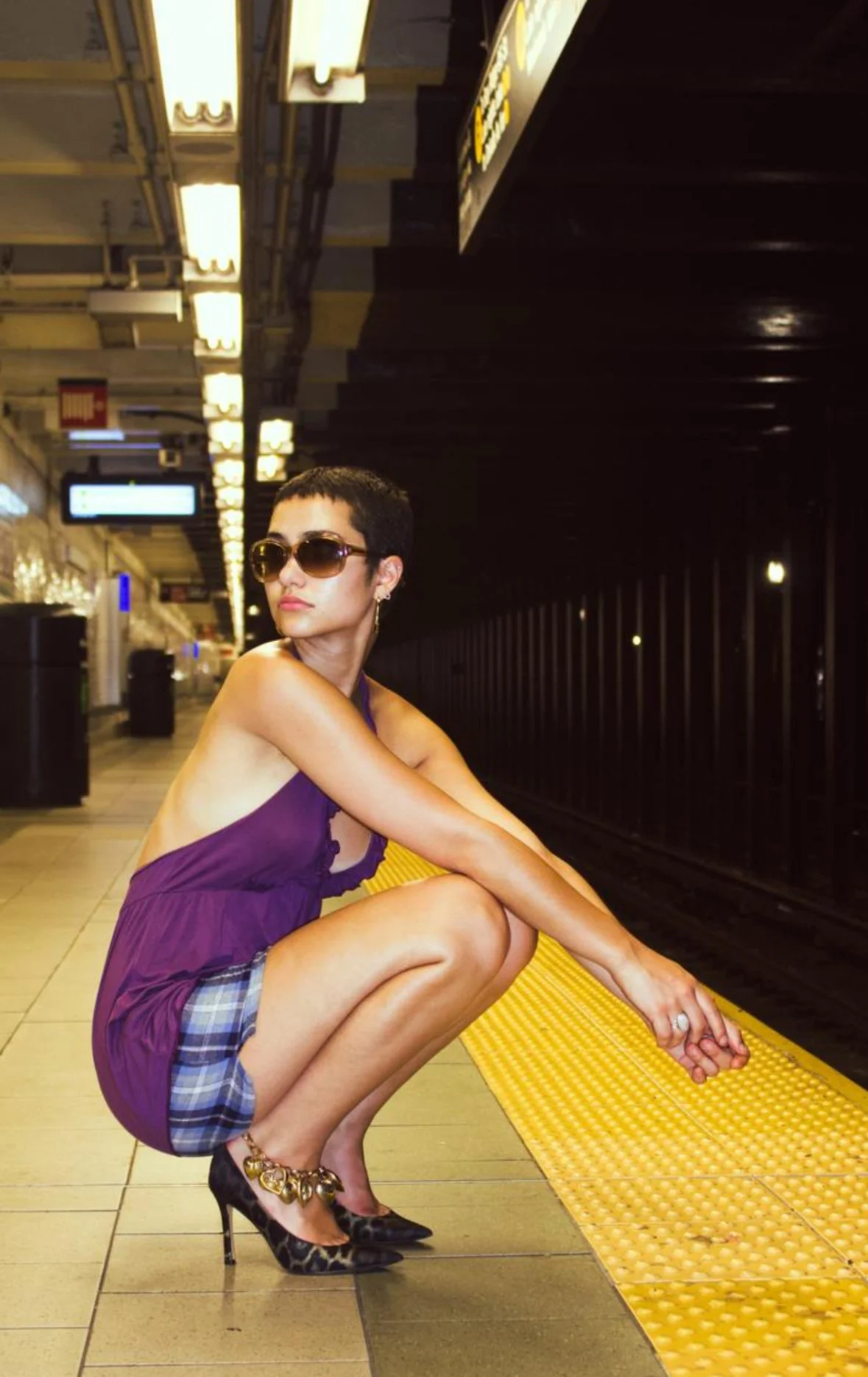 A woman with short dark hair wearing sunglasses, a purple halter top, plaid shorts, and high heels with gold embellishments, squatting on a subway platform near the yellow safety line, with a dark subway tunnel in the background and yellow-lit ceiling lights.