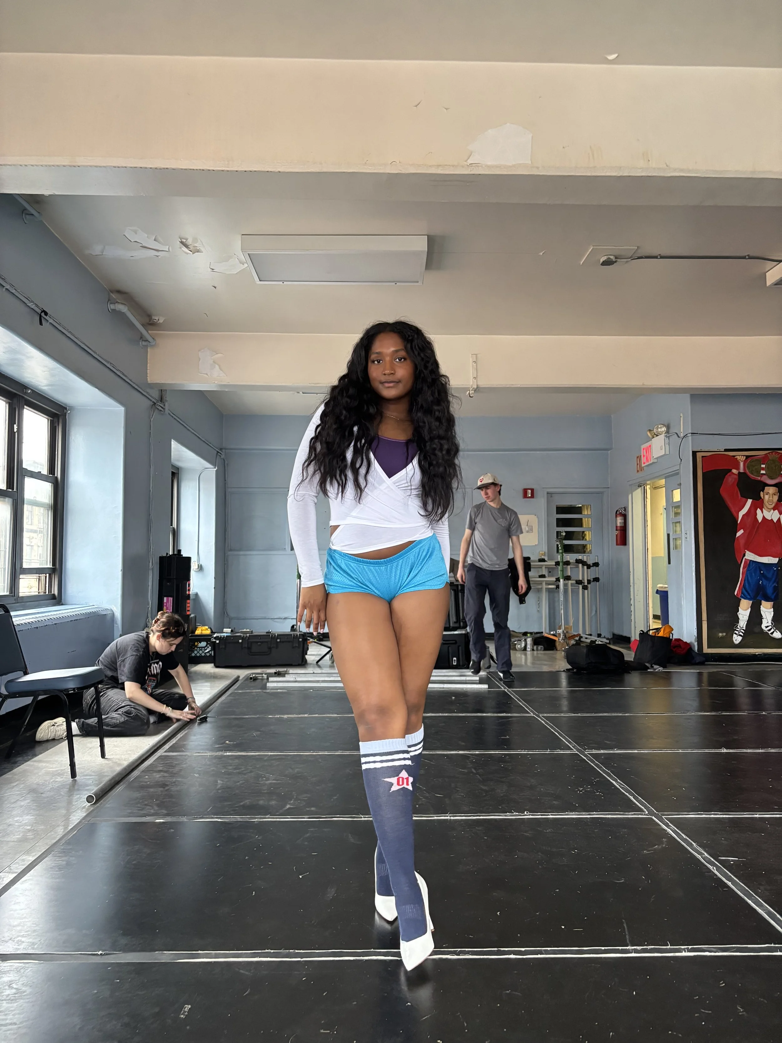 Young woman walking on a black stage in a dance studio, wearing a white long sleeve top, purple shorts, knee-high socks, and white shoes, with a person working on equipment in the background.