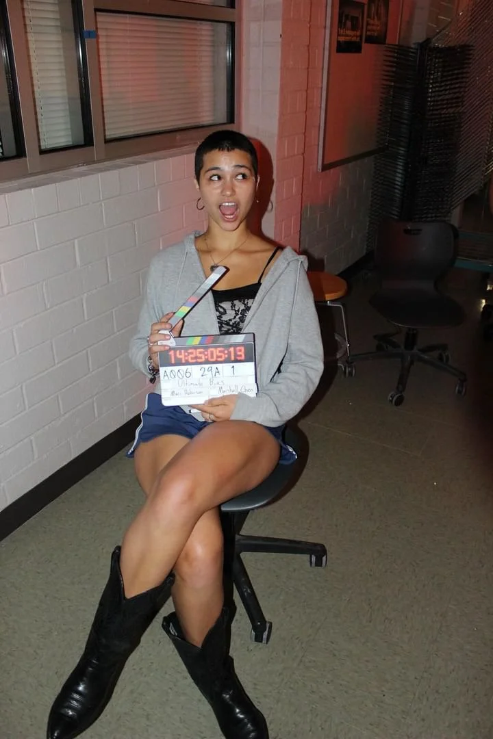 Young woman with short hair and hoop earrings sitting on a swivel chair, holding a clapperboard and a phone, with a surprised expression. She is wearing a gray hoodie, black shorts, and black boots, in an indoor setting with brick walls and windows.