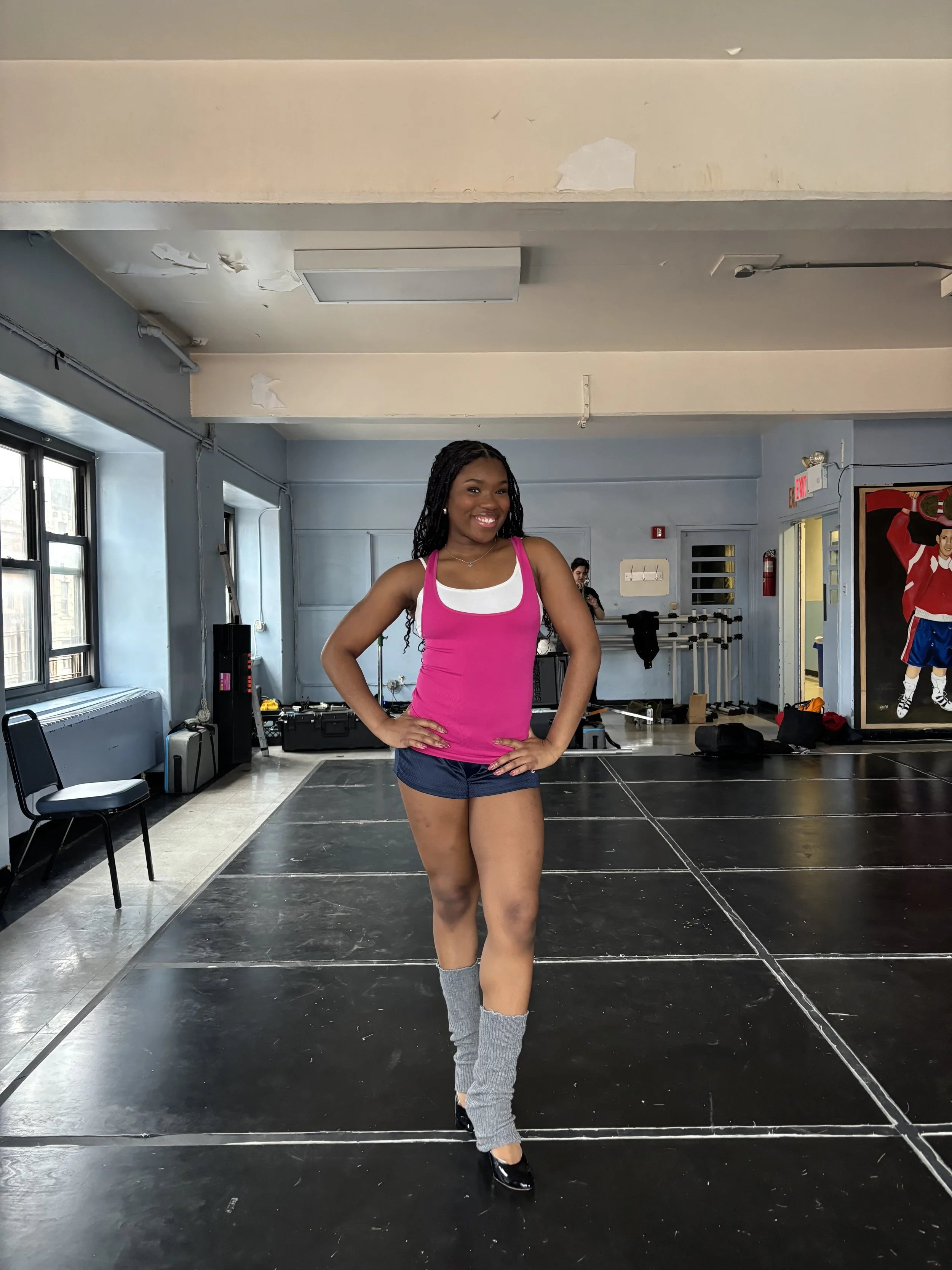 A young woman standing on a dance studio floor, smiling, wearing a pink tank top, navy shorts, gray leg warmers, and black dance shoes.
