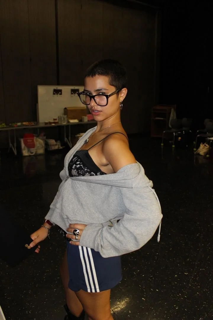 A young woman with short dark hair, wearing glasses, a black lace bralette, a gray hoodie off her shoulders, and navy athletic shorts with white stripes, standing in a dimly lit room.