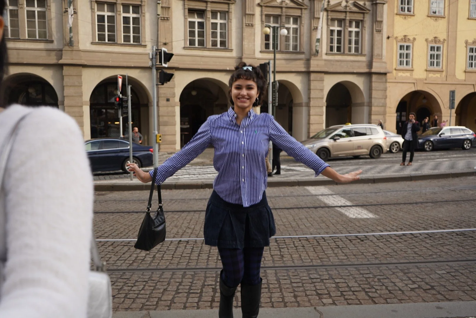 Young woman smiling, standing with arms outstretched on city street near tram tracks, wearing a striped blue shirt, skirt, and rain boots, with cars and pedestrians in the background.