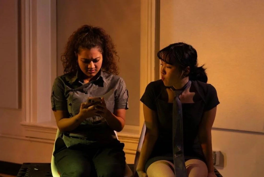 Two women sitting on a bench, one is looking at her phone with a concerned expression, and the other is whispering to her with a serious look, in a dimly lit room.