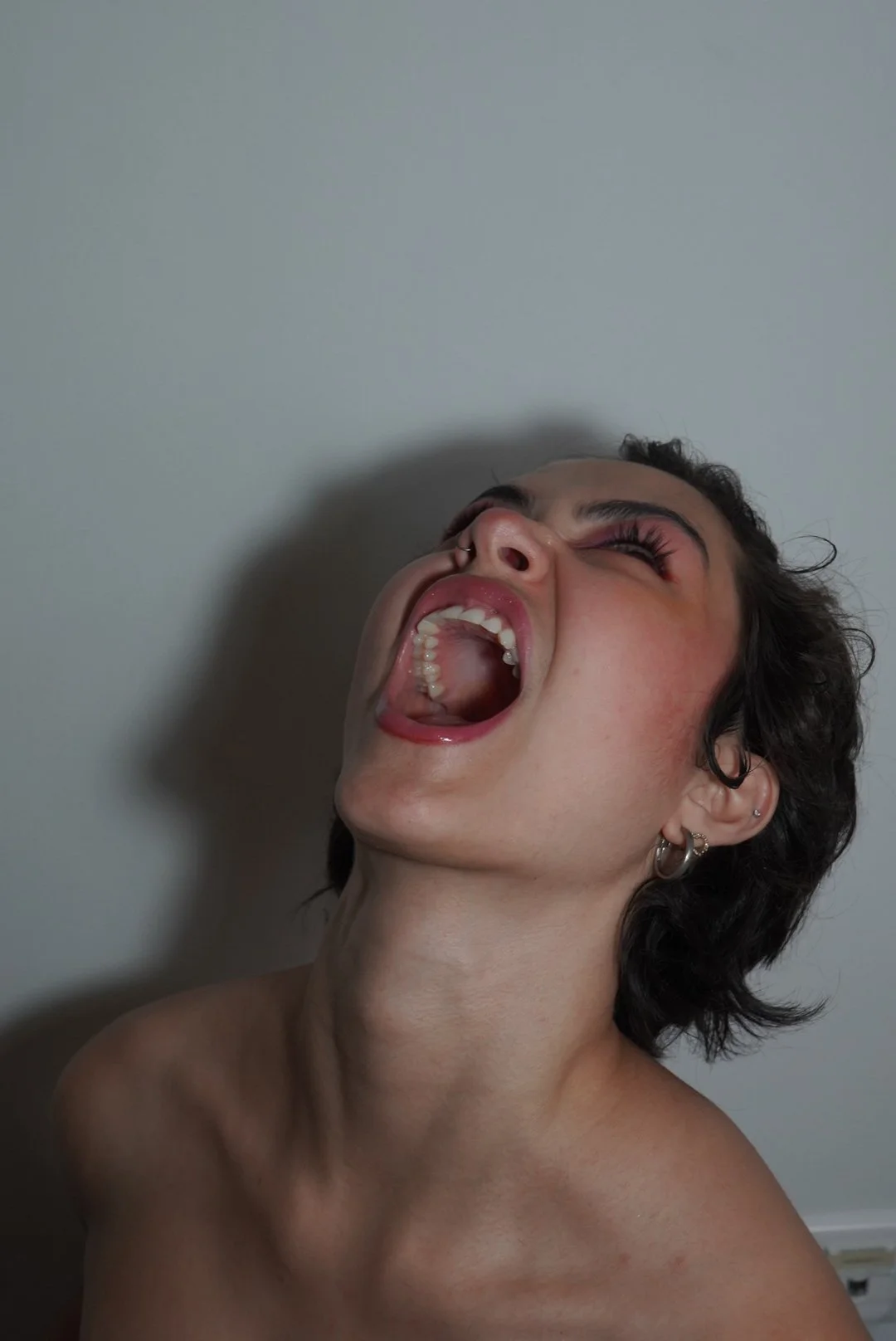 A person with short dark curly hair, multiple earrings, eyes closed, and mouth open wide, appearing to scream or shout upward against a plain wall background.