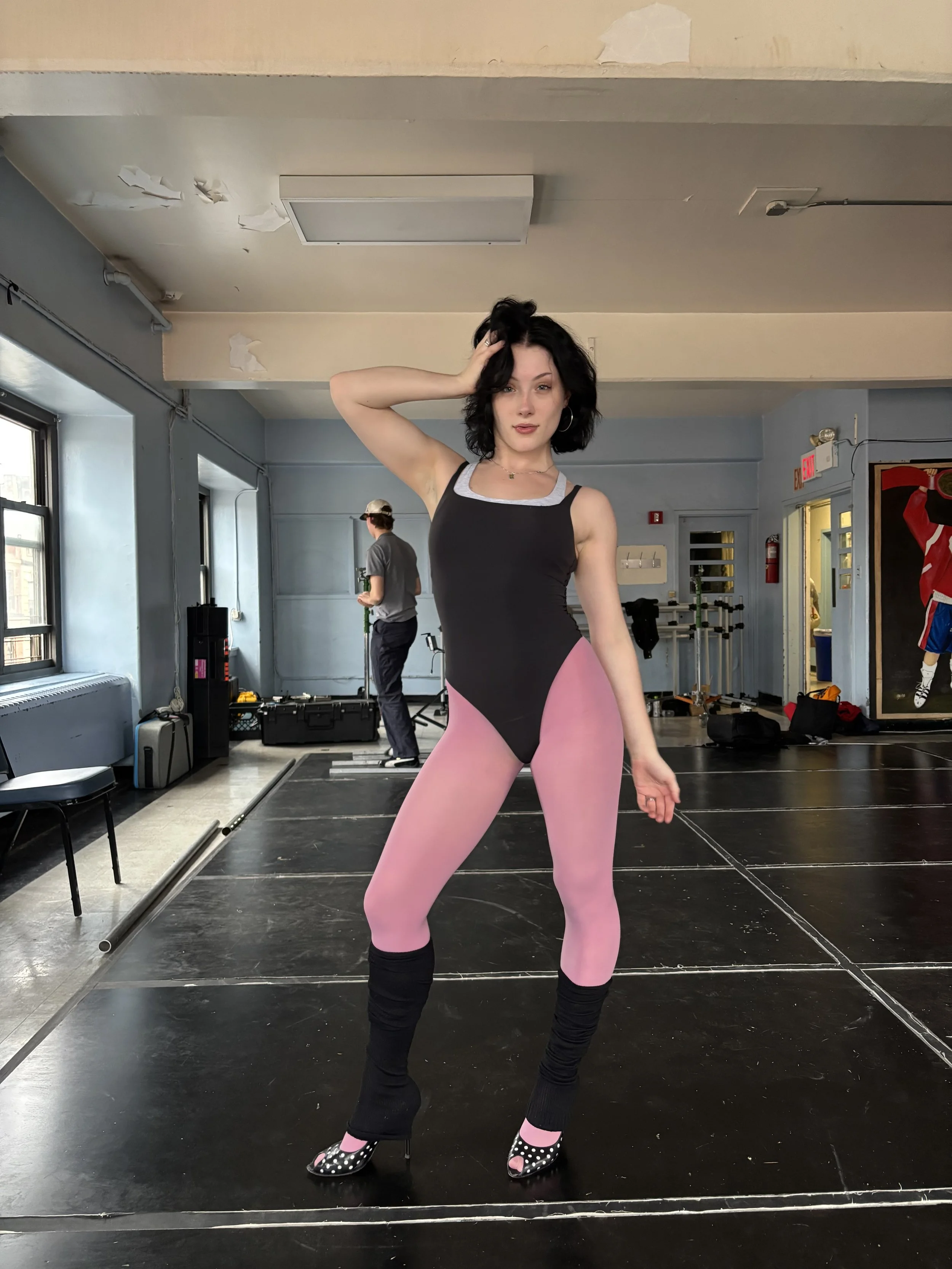 A woman with black hair in a dance studio, wearing a black and grey bodysuit, pink tights, and black high-heeled shoes with white polka dots.