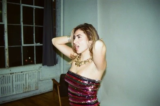 Singer Songwriter Karina Eleni in a strapless sequin dress with wavy hair posing indoors near a window.