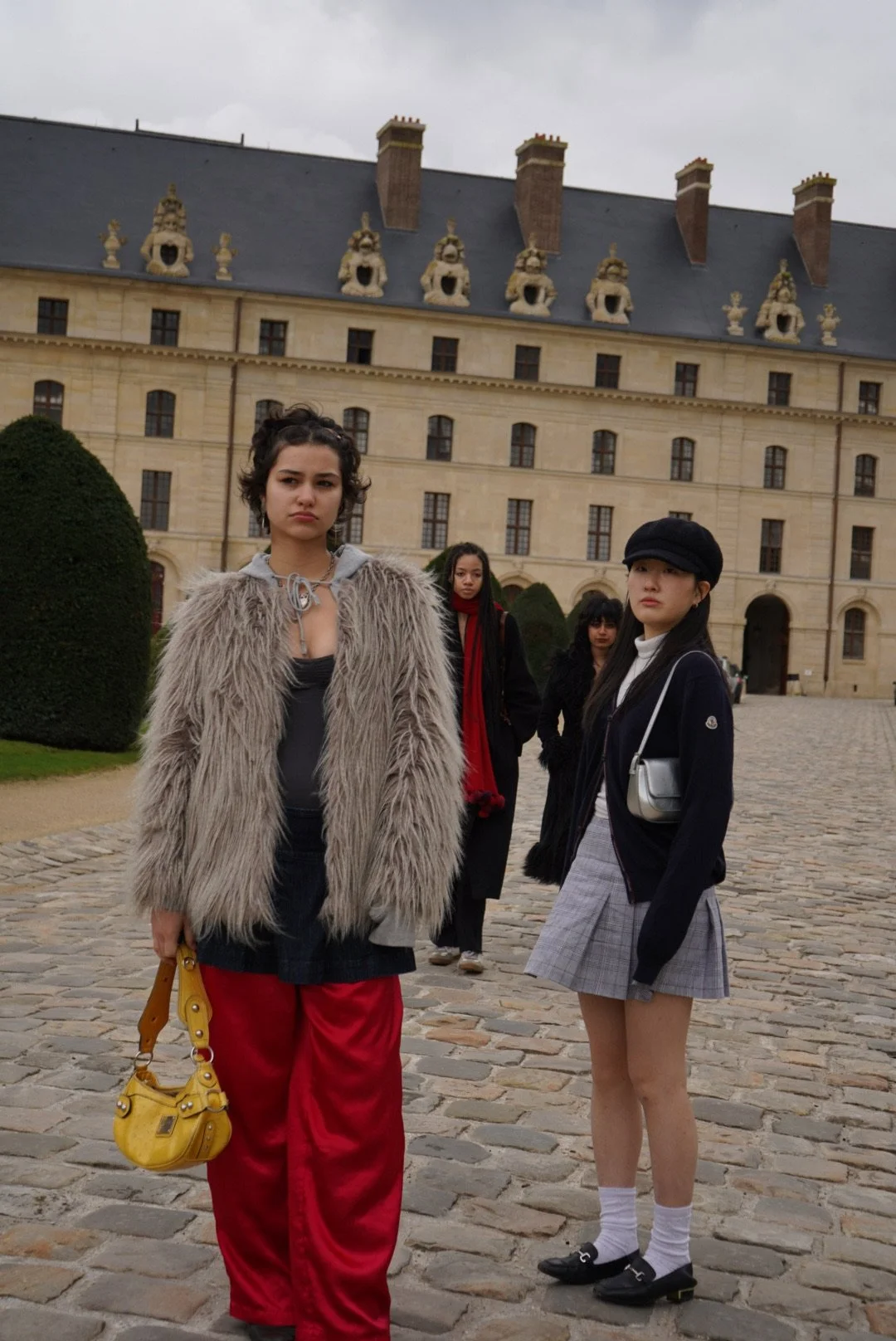 Four young women standing on a cobblestone courtyard in front of a historic, castle-like building with ornate rooftop sculptures. Two women in the foreground look at the camera; one wears red baggy pants and a fur coat, and the other wears a gray plaid skirt, black jacket, and black cap.