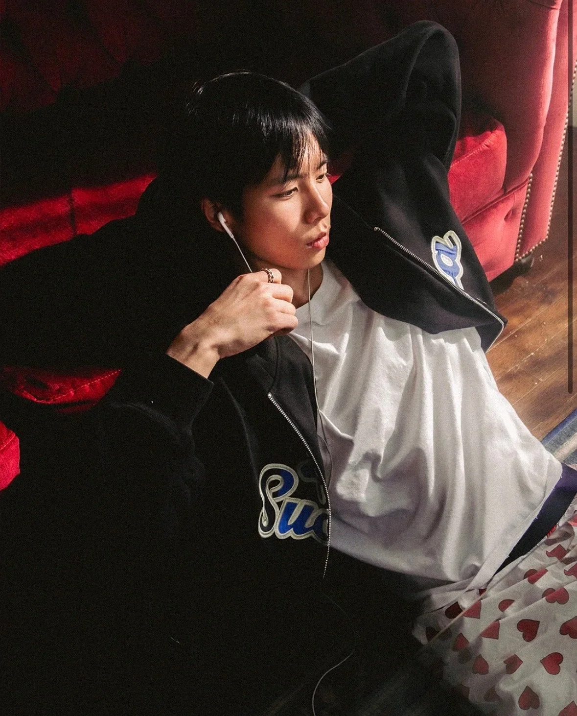 A young man with black hair is lying on a red couch, listening to music with white earbuds. He is wearing a black jacket with a patch and a white shirt underneath. His right hand is near his face, and his left arm is resting behind his head. He appears to be relaxed, with a soft expression on his face.