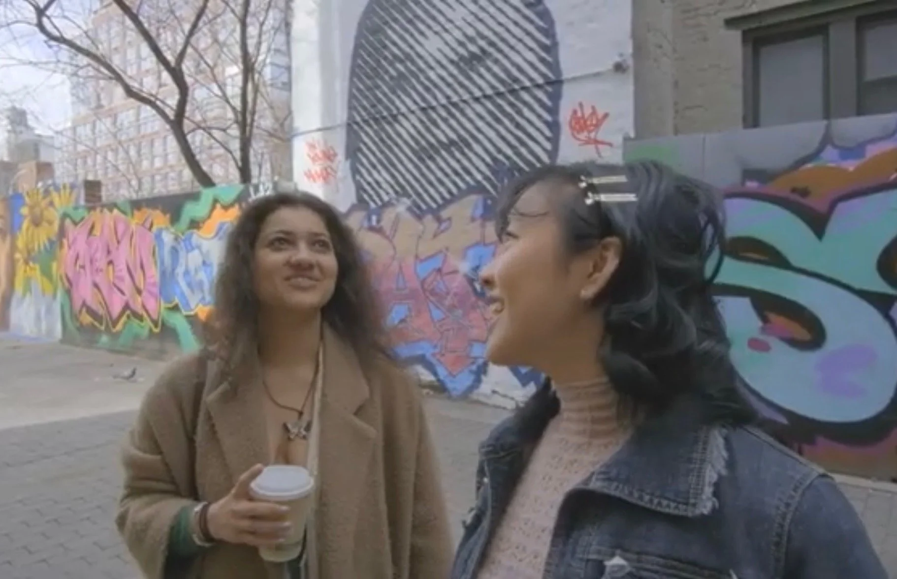 Two women smiling and talking outdoors on a city street with colorful graffiti on a wall behind them.