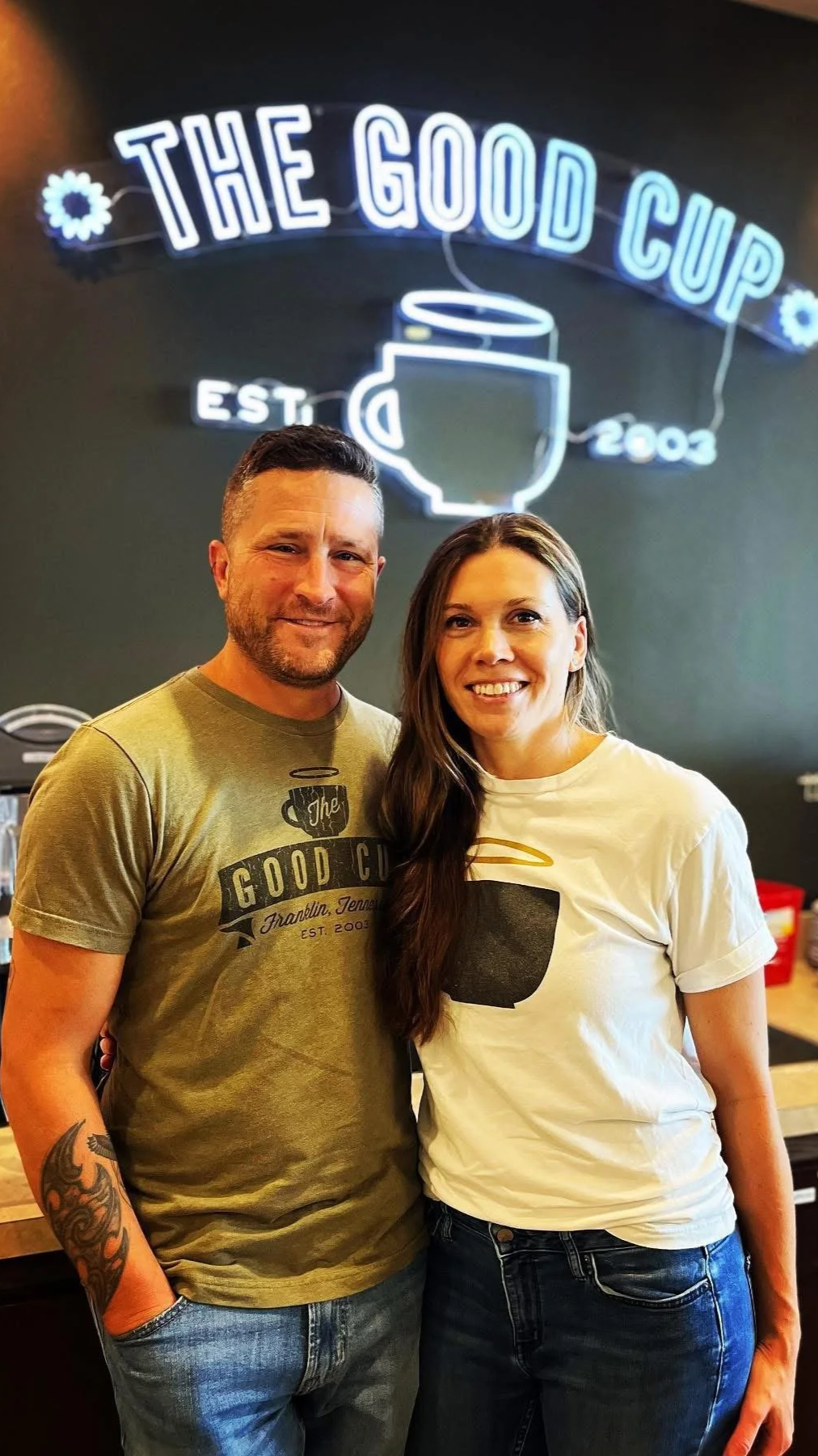 A man and woman standing inside a cafe called The Good Cup, with a neon sign of a coffee mug and the cafe's name in the background.