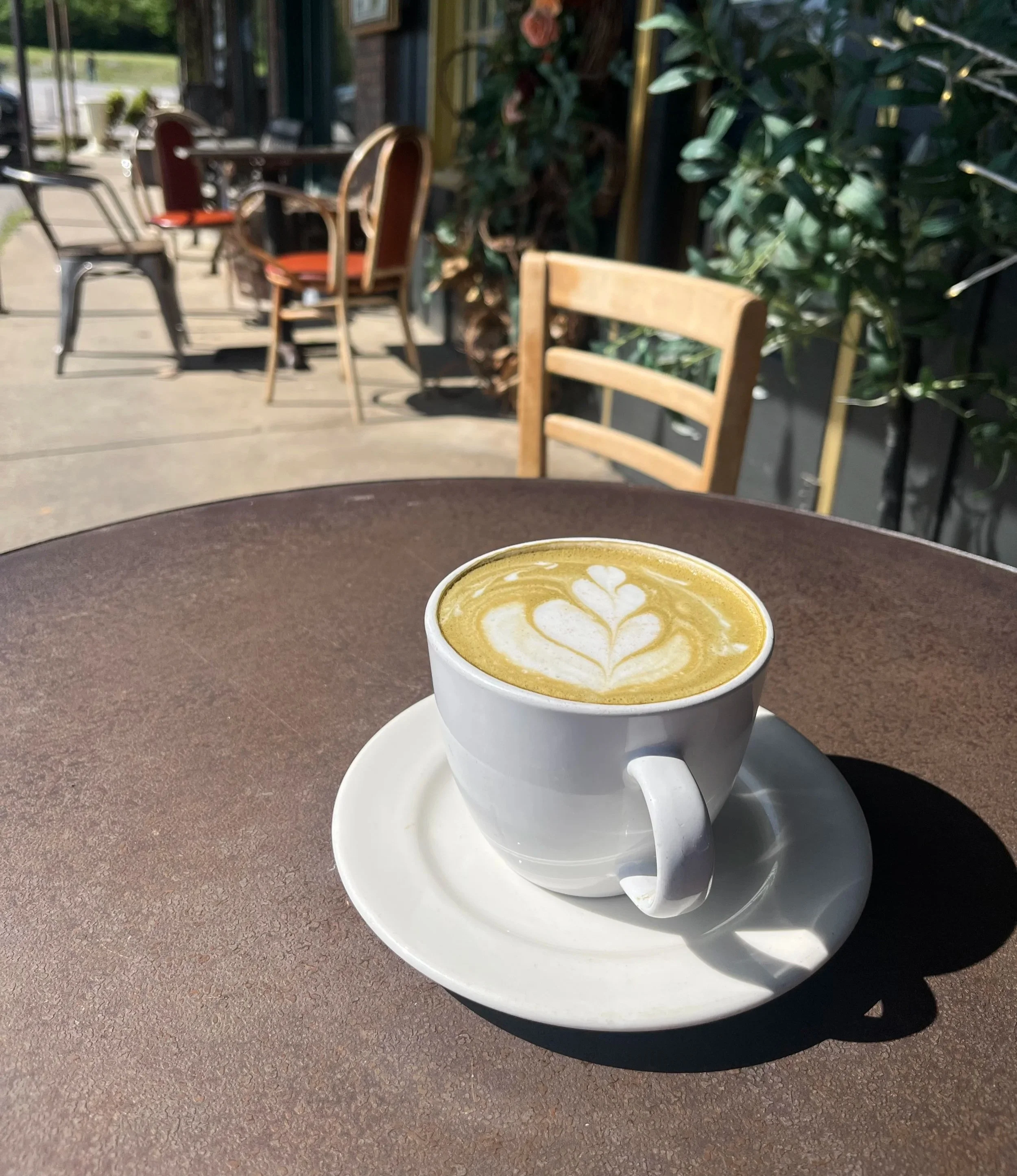 A white cup of matcha latte with latte art on top, sitting on a white saucer on a brown outdoor table at a cafe, with chairs and greenery in the background.