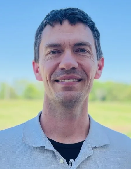 A man smiling outdoors with a blue sky and green trees in the background.