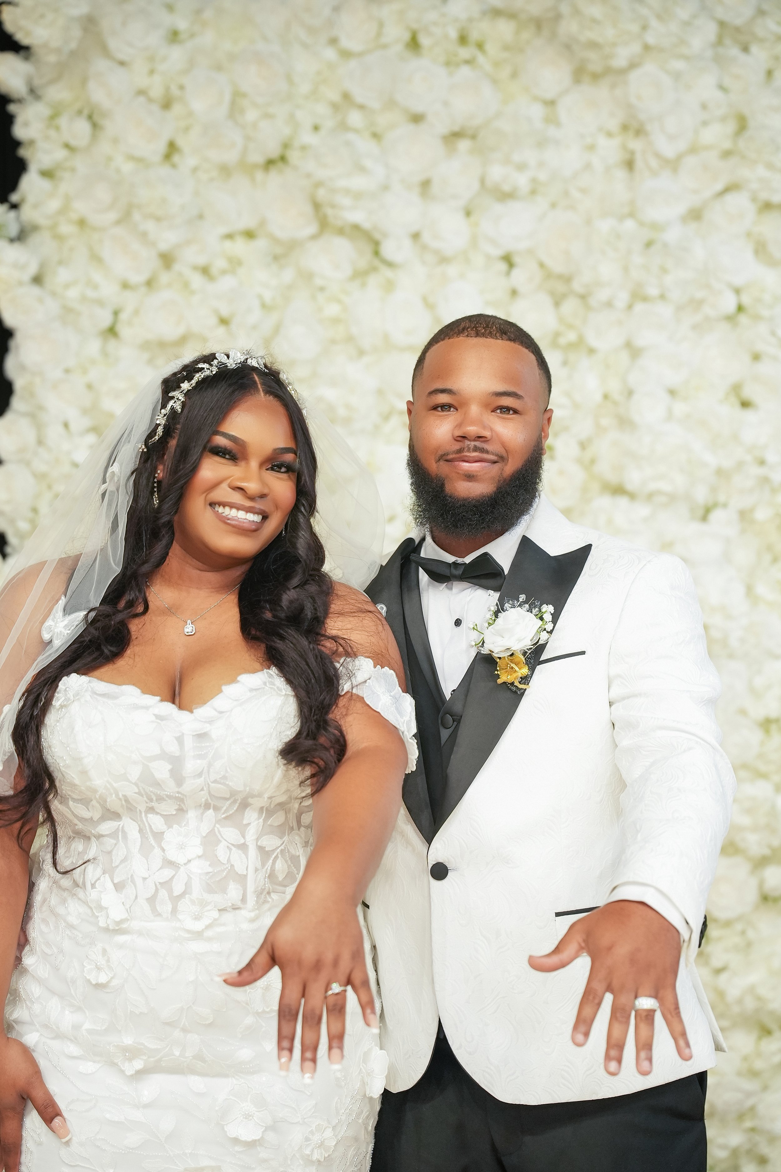 A bride and groom display their wedding rings in front of a white floral backdrop.