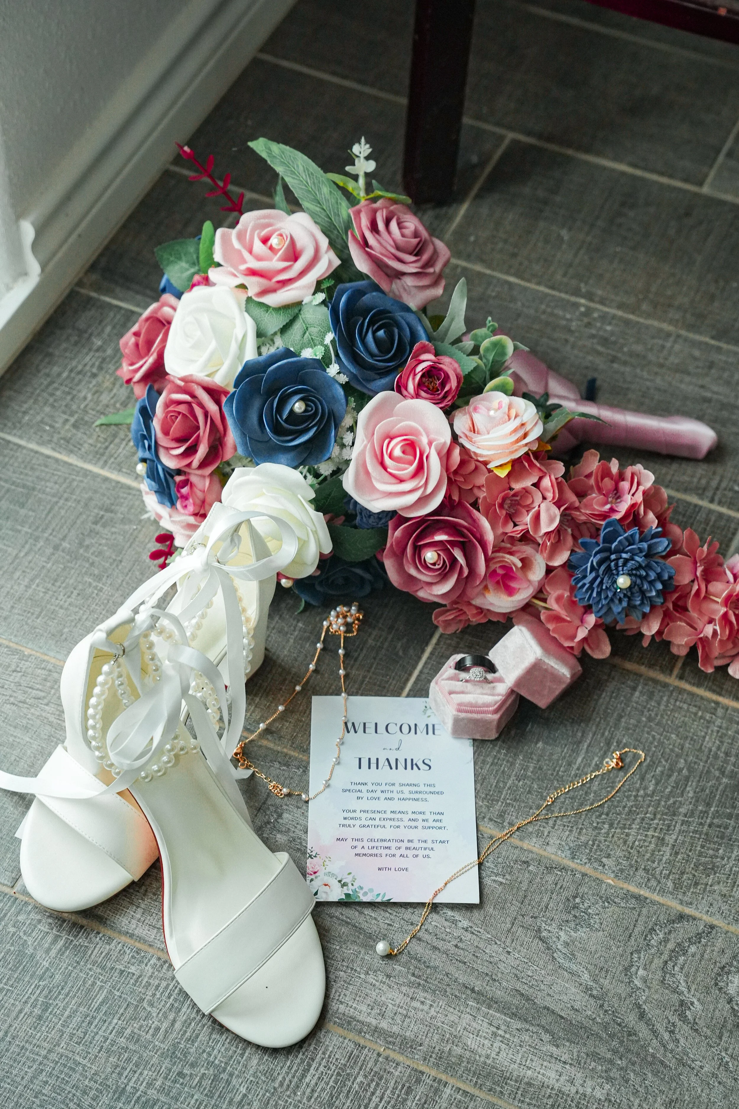 A bouquet of pink, white, blue, and purple artificial roses and hydrangeas, a pair of white high-heeled shoes, a greeting card that says "Welcome and Thanks," a gold necklace with pearls, a pair of pearl earrings, and a small pink case with a ring inside, all placed on a tiled floor.