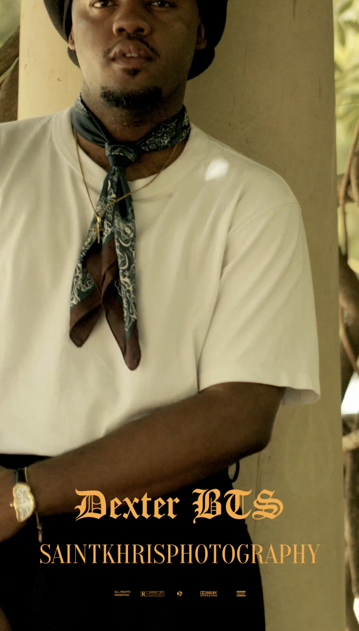 Close-up of a young man wearing a white t-shirt, a patterned scarf, a black hat, and a gold watch, standing near a wall with a blurred background.