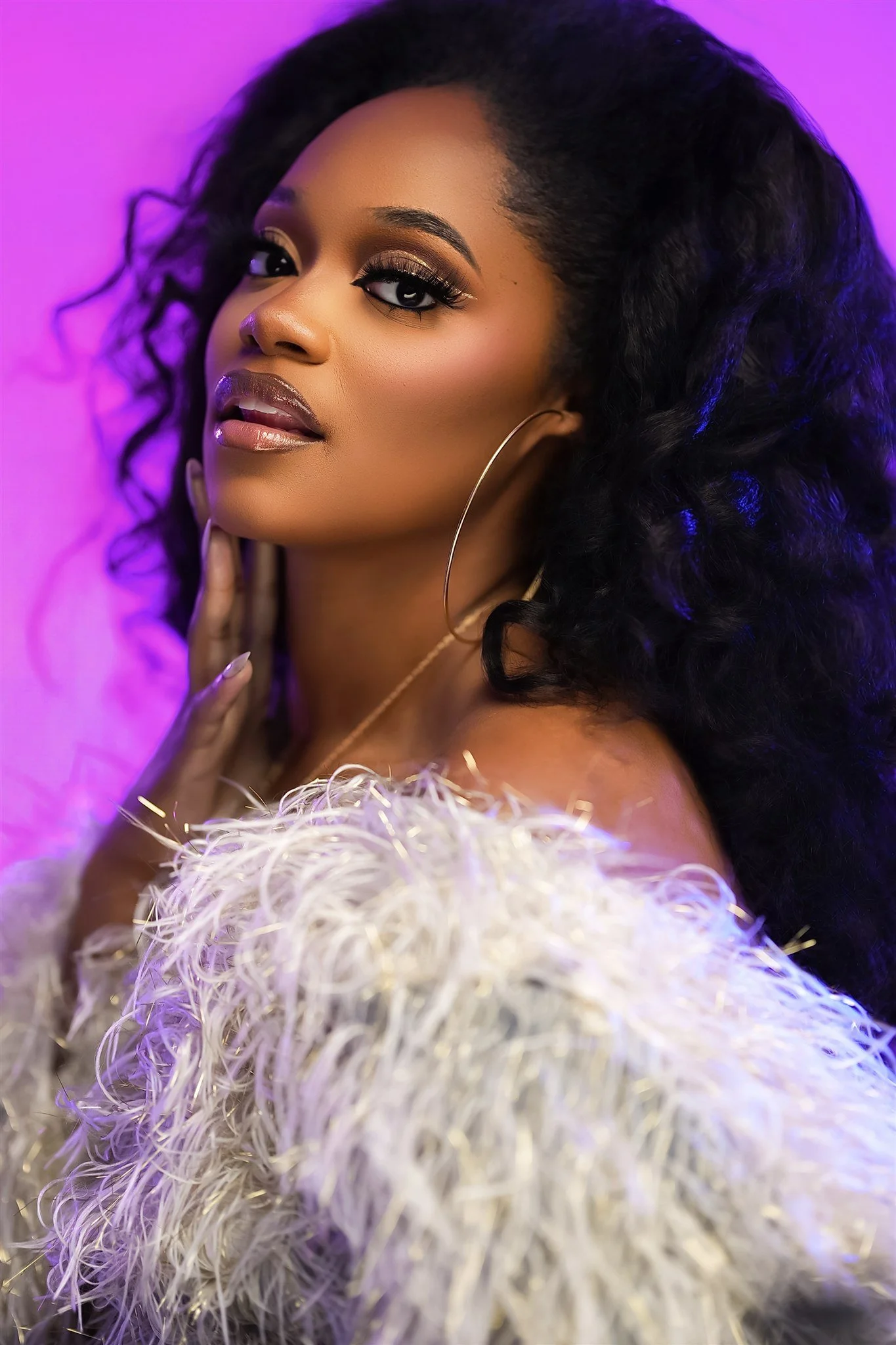 A woman with curly black hair and makeup, wearing a white feathered outfit and large hoop earrings, posing against a purple background.