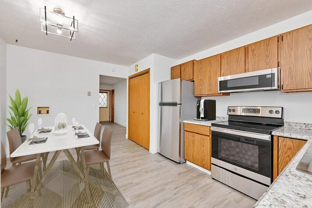 A kitchen and dining area with a white table, six brown chairs, wooden cabinets, a stainless steel refrigerator, a microwave, and a stove, with a ceiling light fixture.