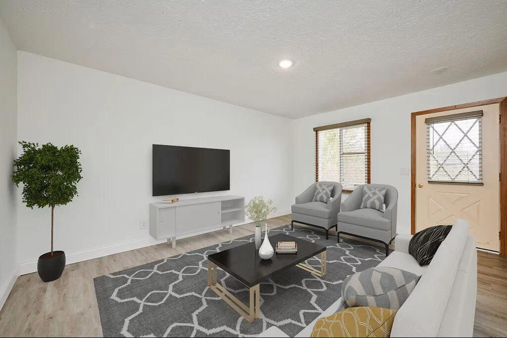 Living room with white walls, a flat-screen TV on a white TV stand, a gray patterned rug, two gray armchairs, a white sofa with patterned pillows, a black coffee table with decorative items, a window with wooden blinds, and a door with metal bars, all lit by natural and ceiling light.