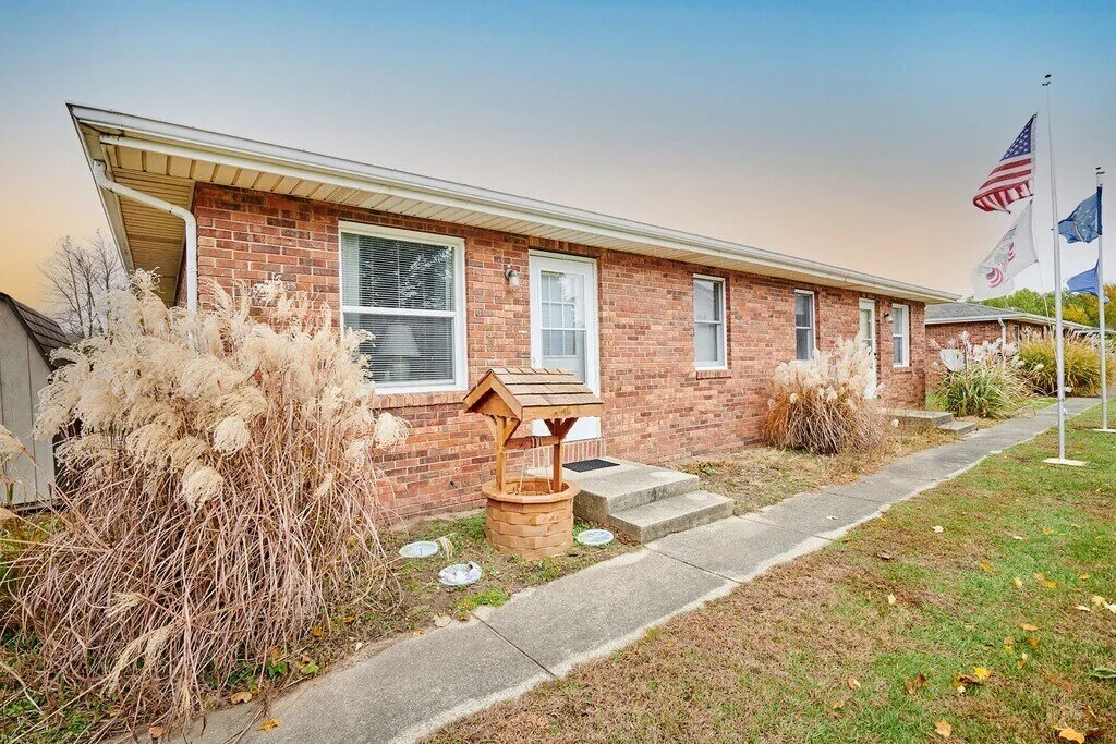 Brick house with a slanted roof, three front windows, and a white door. There are three flags on poles and decorative grasses in the yard.