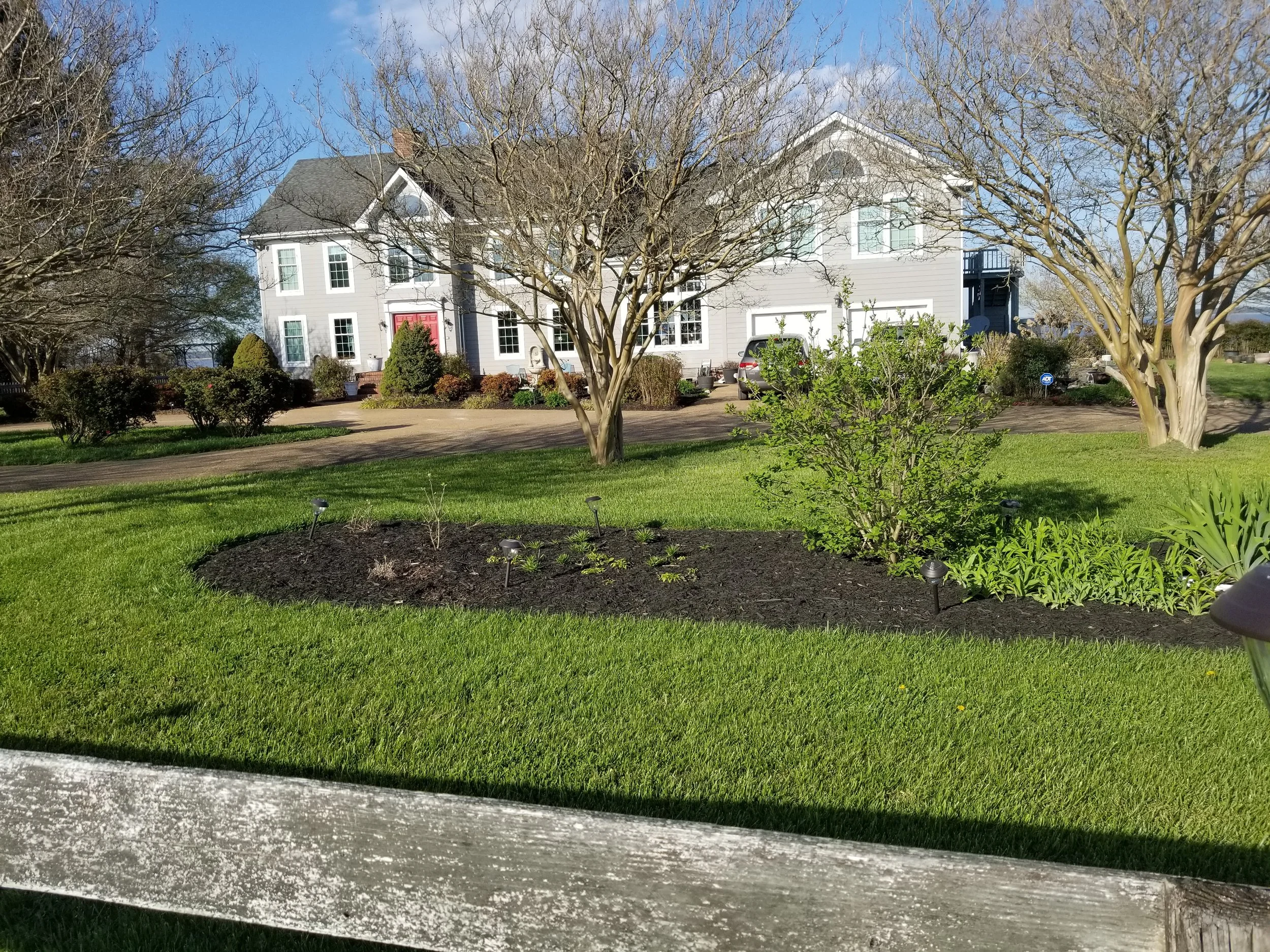 A large, two-story house with a white exterior and red front door, surrounded by grass, trees, and garden beds, with a driveway and parked cars.