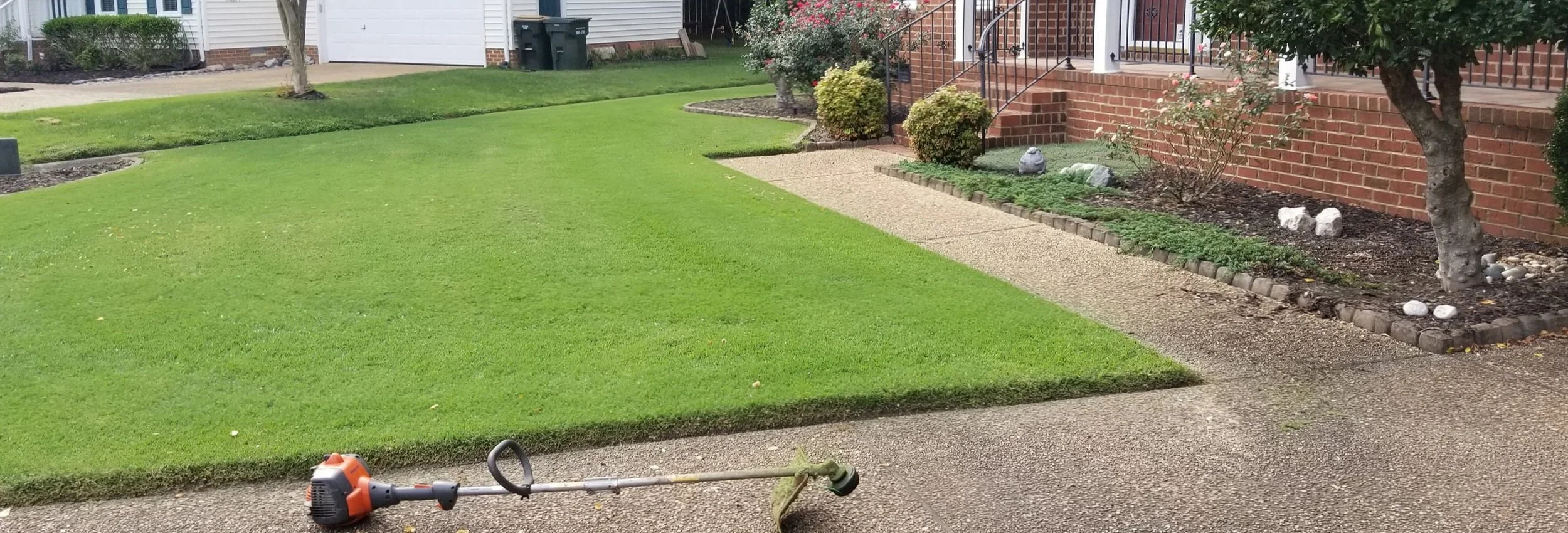 A freshly mowed front yard with green grass, a walkway, and a brick house with stairs and plants.