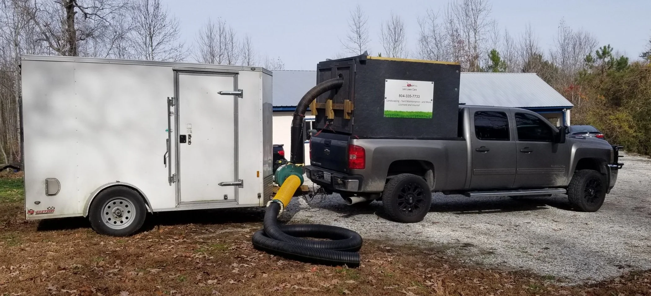 A gray pickup truck attached to a white enclosed trailer and a black wood chipper with a large flexible hose connected. The scene is outdoors with trees and a building with a metal roof in the background.