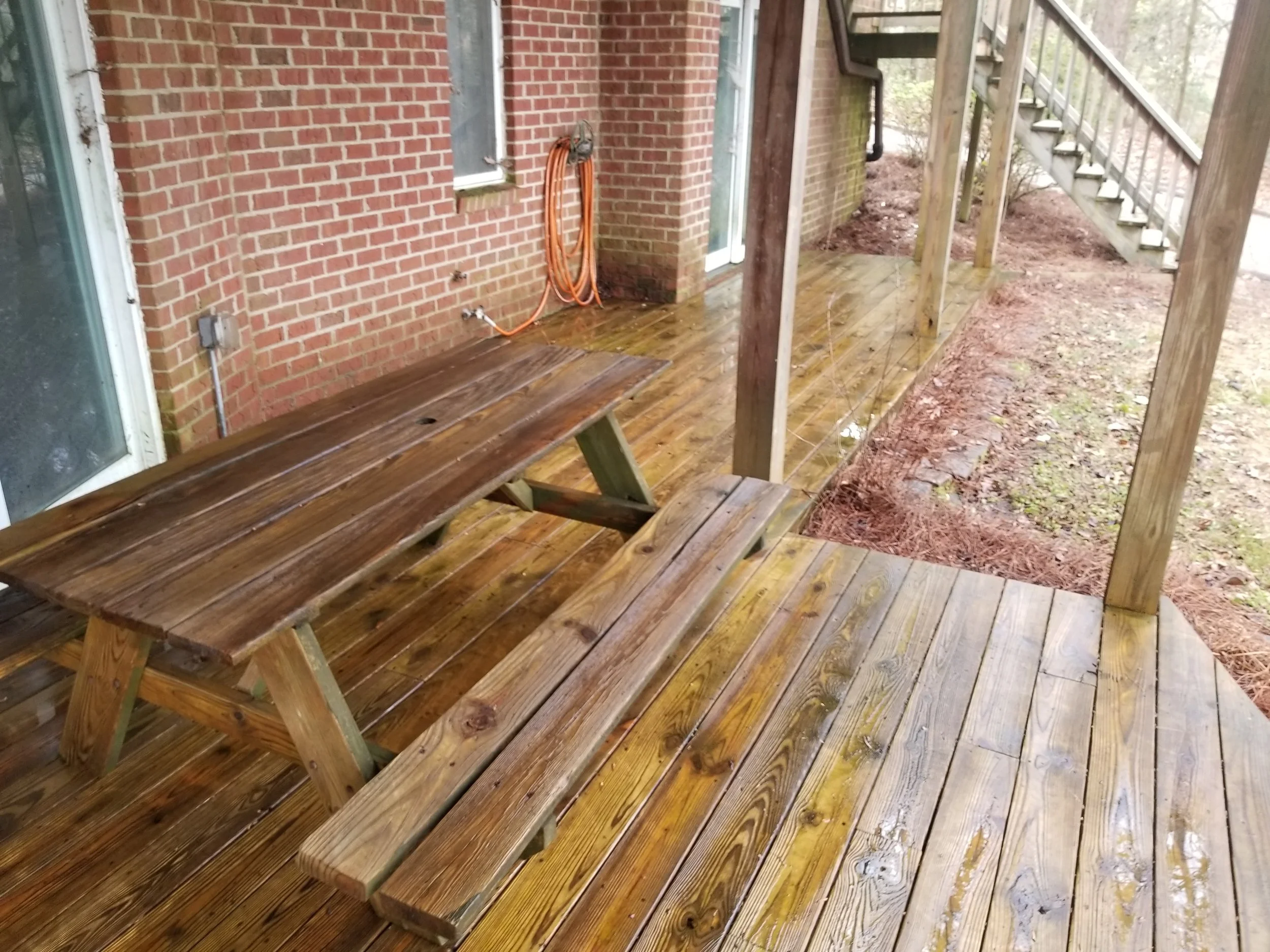 A wet wooden deck with a picnic table attached to a brick house, and stairs leading up to an outdoor area.
