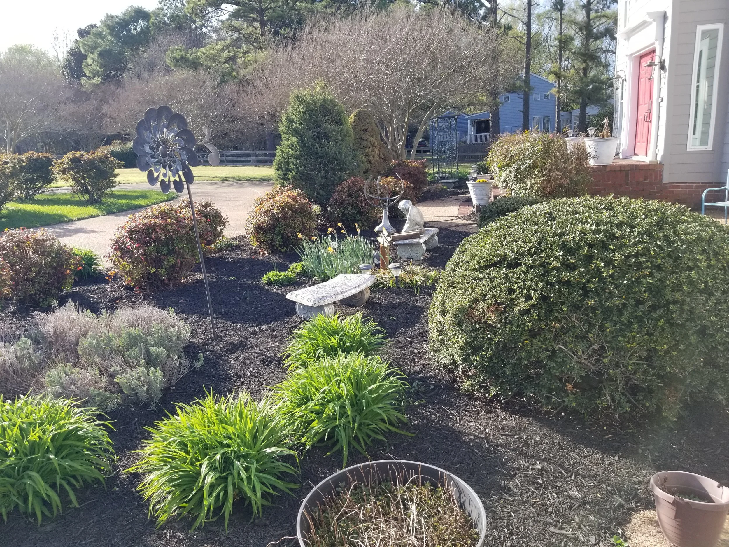 A landscaped garden with various bushes, plants, and decorative objects such as a metal flower wind spinner, a fountain, small benches, and ornamental statues. The garden is adjacent to a house with a red door and windows, with neighboring houses visible in the background.
