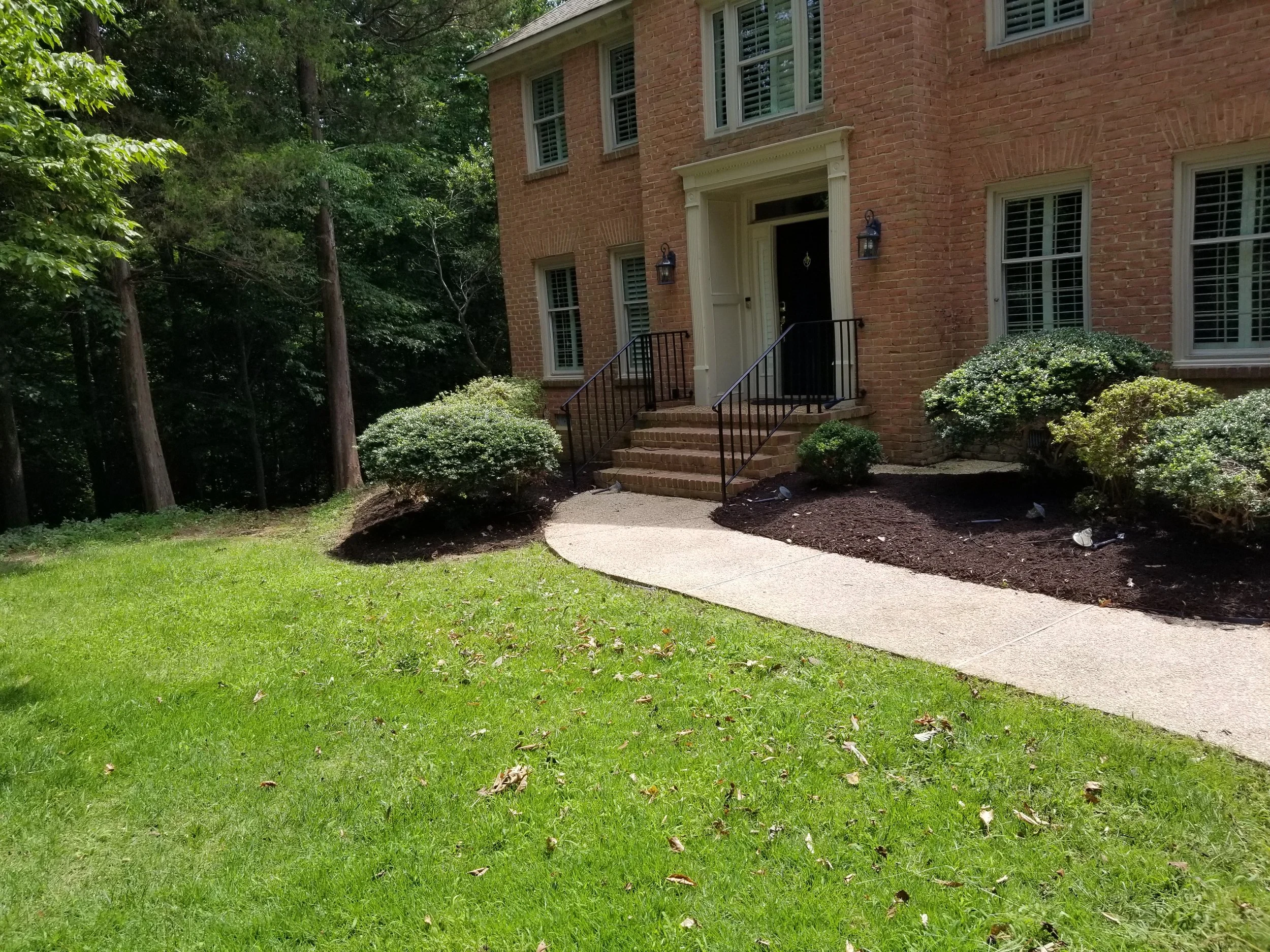Front of a brick house with steps leading to a black door and black handrails. Well-maintained lawn with small bushes and a winding concrete walkway. Tall trees are visible in the background.