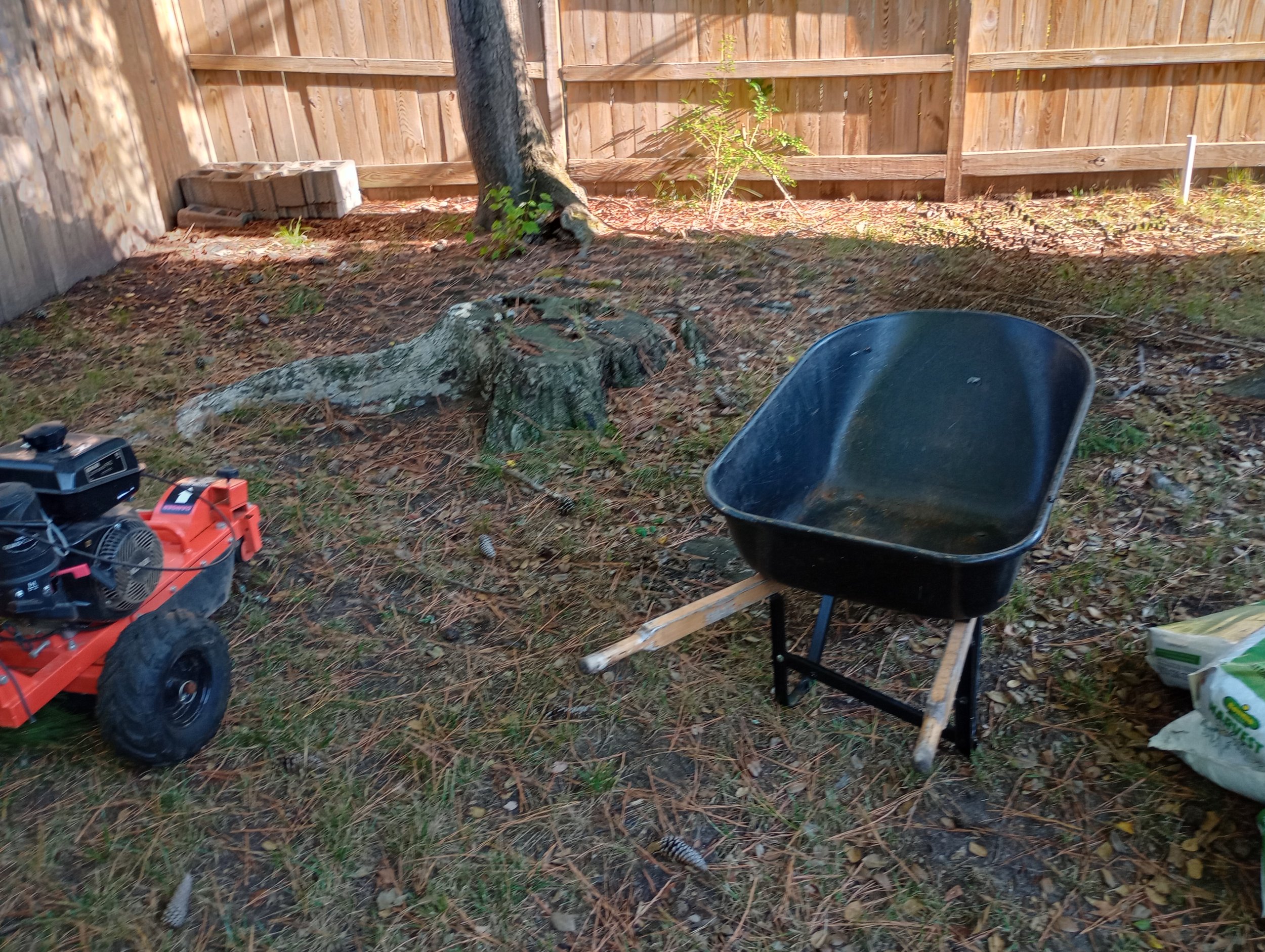 Backyard with a wheelbarrow, leaf blower, tree stump, and wood fence.