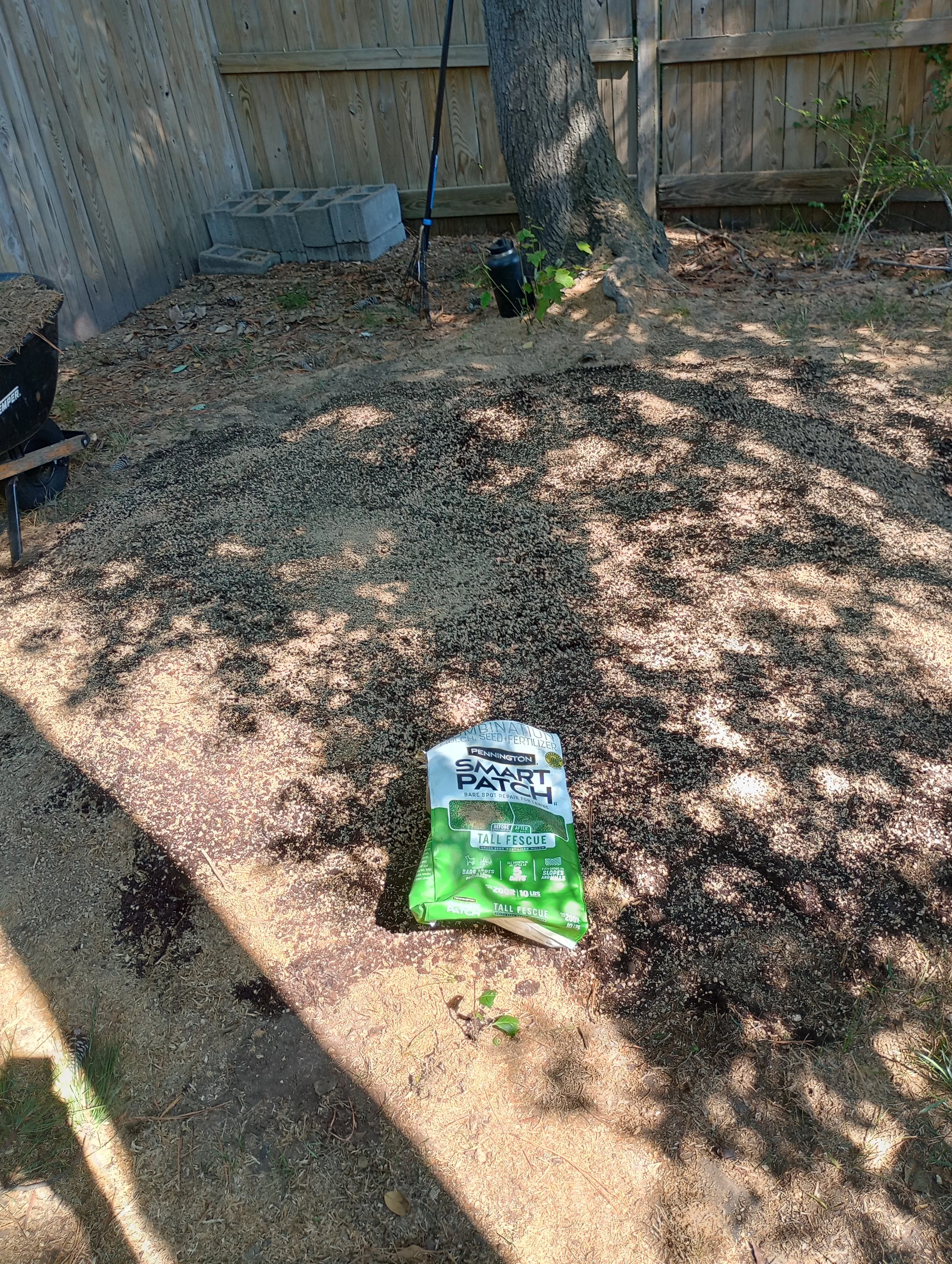 Bag of trill seed fertilizer placed on a dirt area in a backyard, surrounded by a wooden fence, a tree, and gardening tools like a rake and a water bottle.