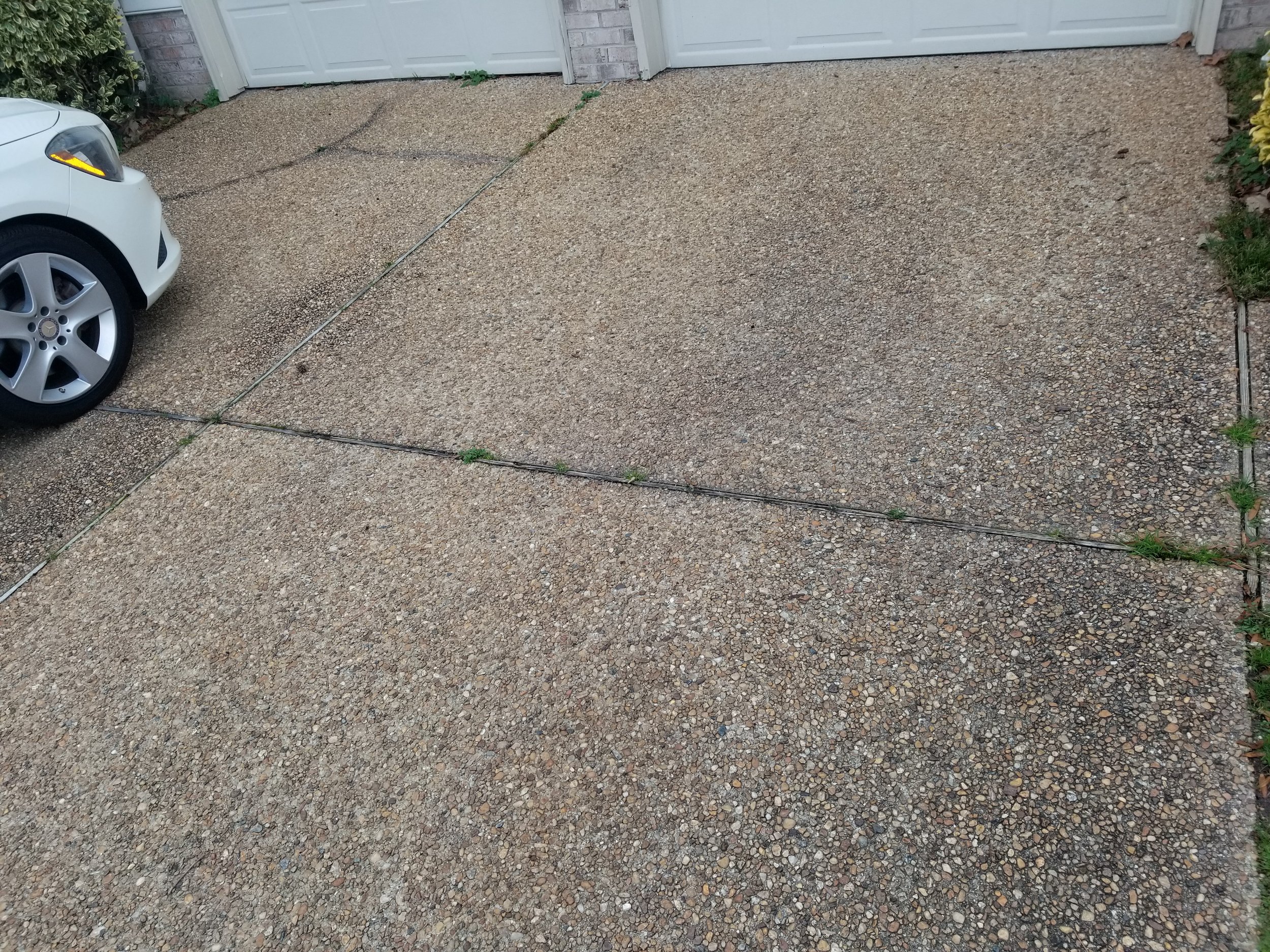 A driveway with small pebbles, a portion of a white car on the left, a garage door in the background, and some green plants on the right side.