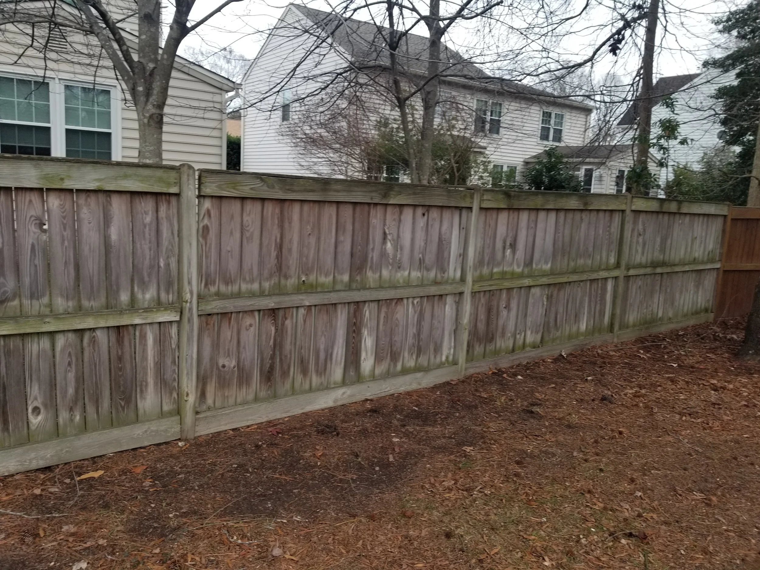 A wooden privacy fence with horizontal panels and vertical posts in a backyard, with houses and trees visible in the background under an overcast sky.