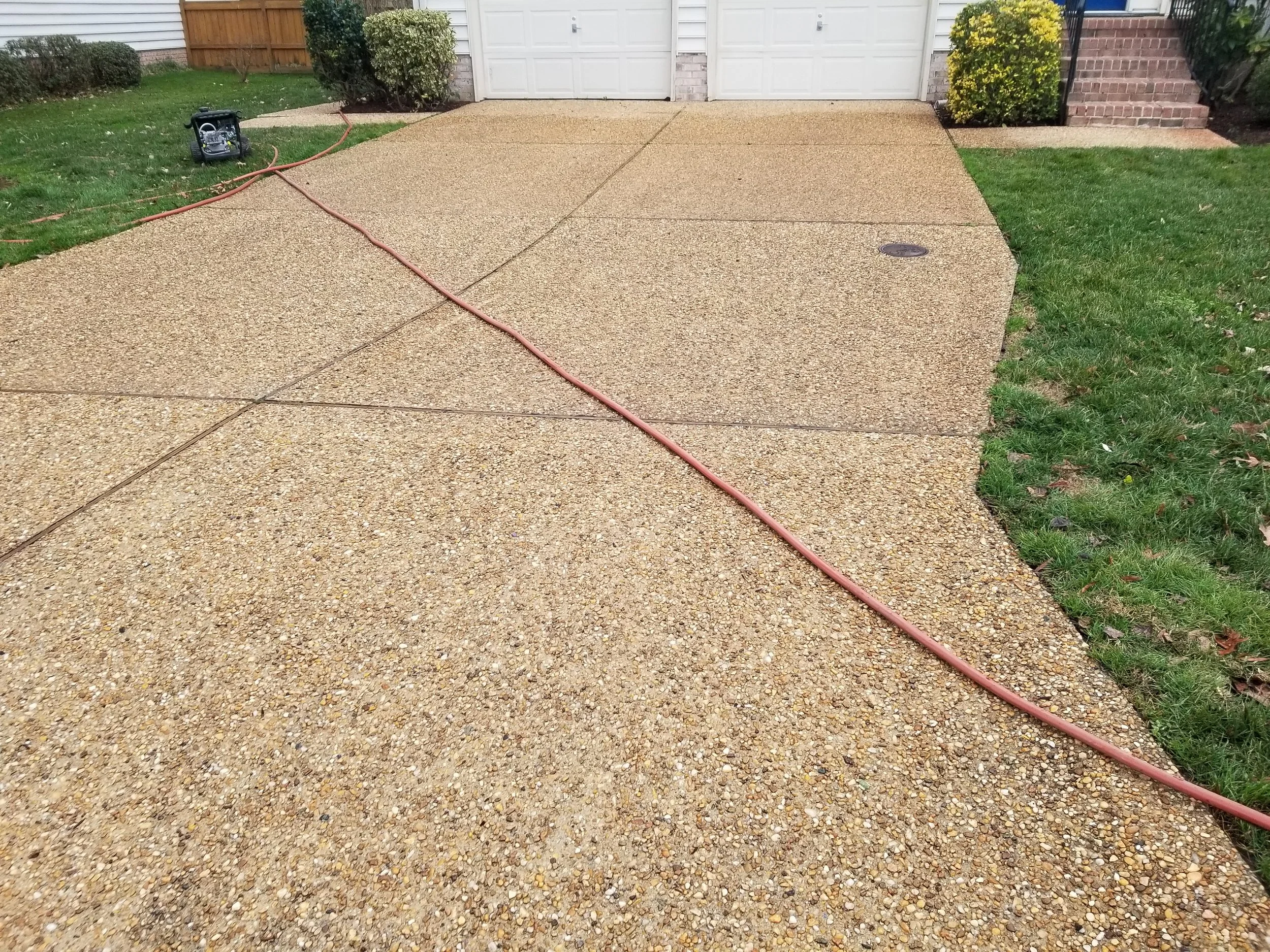 Newly poured concrete driveway with a hose lying on it, leading to an outdoor power generator, in front of a house with white garage doors, surrounded by green grass and shrubs.