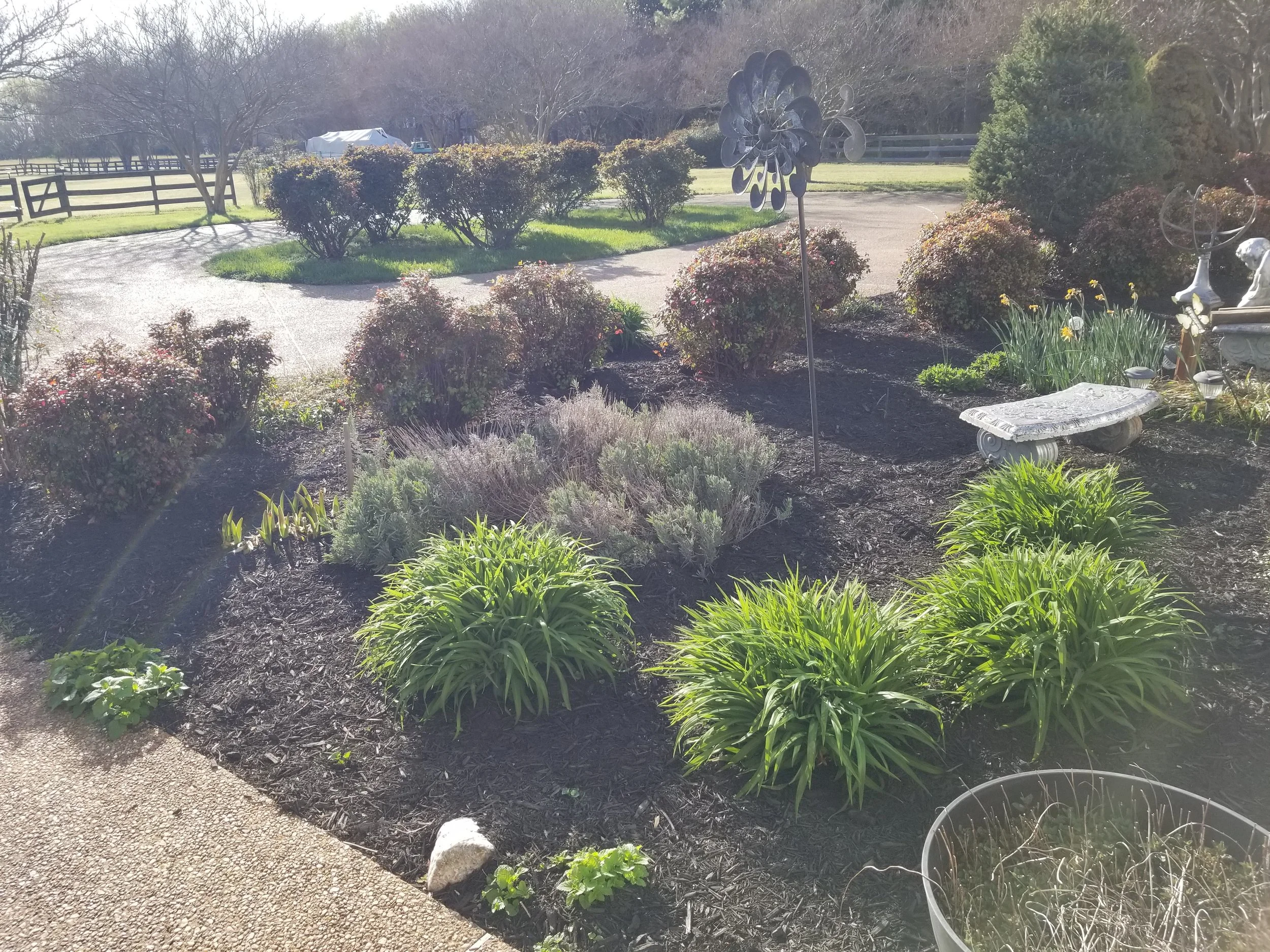 A garden with ornamental bushes, flowering plants, a stone bench, garden sculptures, and decorative weather vanes.
