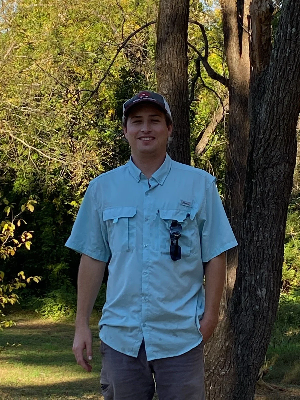 A smiling young man standing outdoors near trees in a park or forested area during daytime, wearing a light blue short-sleeved button-up shirt, sunglasses hanging from his shirt pocket, a baseball cap, and gray shorts.