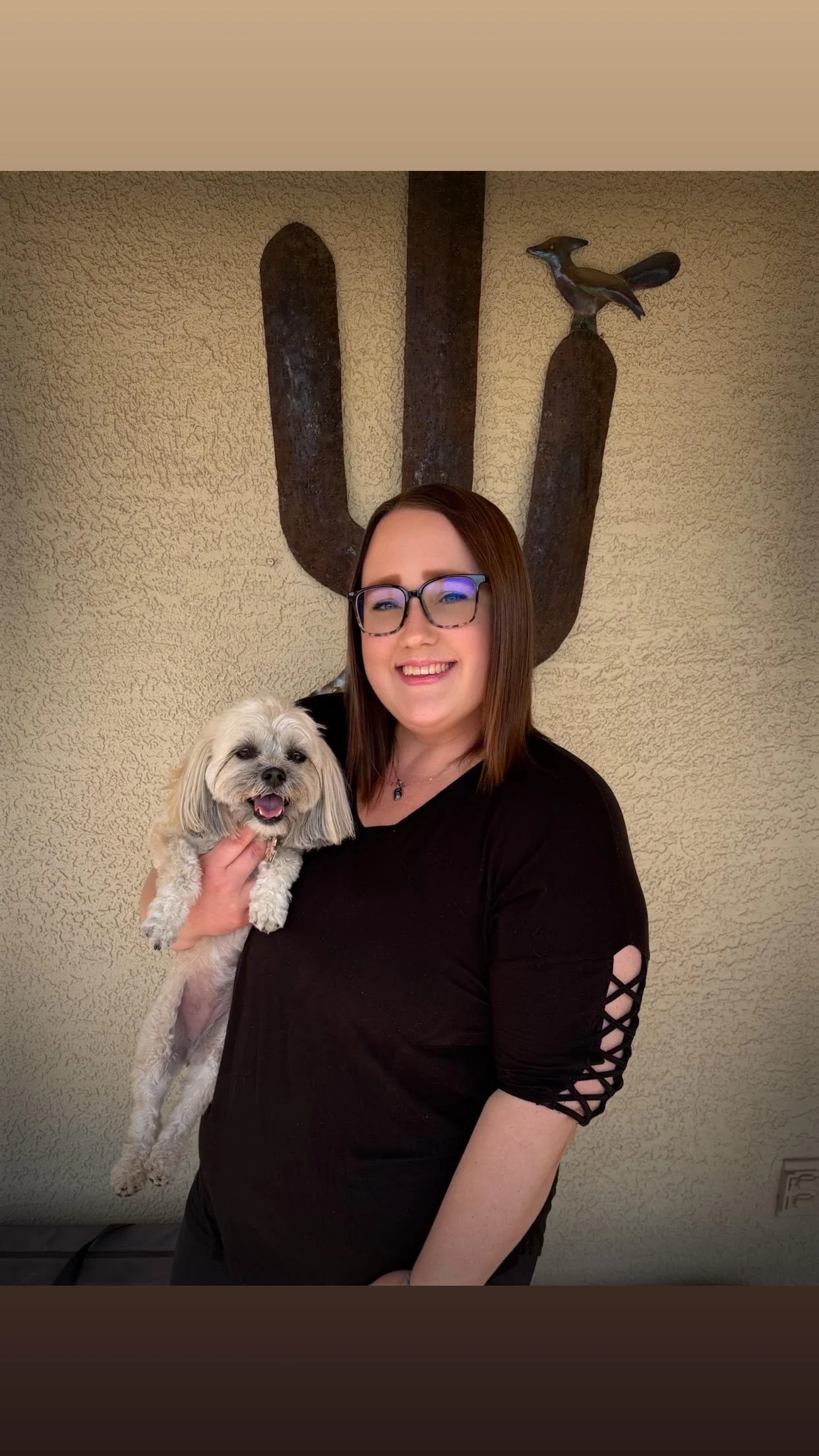A woman with glasses holding a small, fluffy dog with a happy expression standing in front of a textured beige wall with large rust-colored metal western-themed letter and bird decorations.