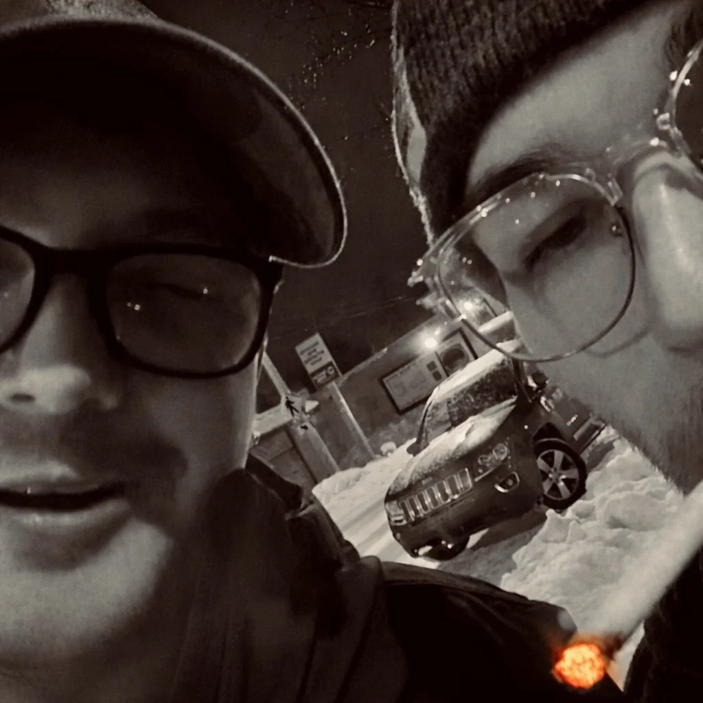 Two young men with glasses and beanies posing close together outdoors in winter, with snow-covered ground and a parked car in the background.