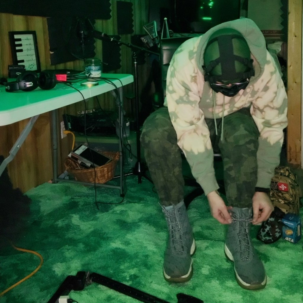 A person dressed in camouflage pants and a hoodie is sitting on a stool, tying the laces of their hiking boots in a music studio or recording space, with soundproofing panels, audio equipment, and a green-painted floor around them.