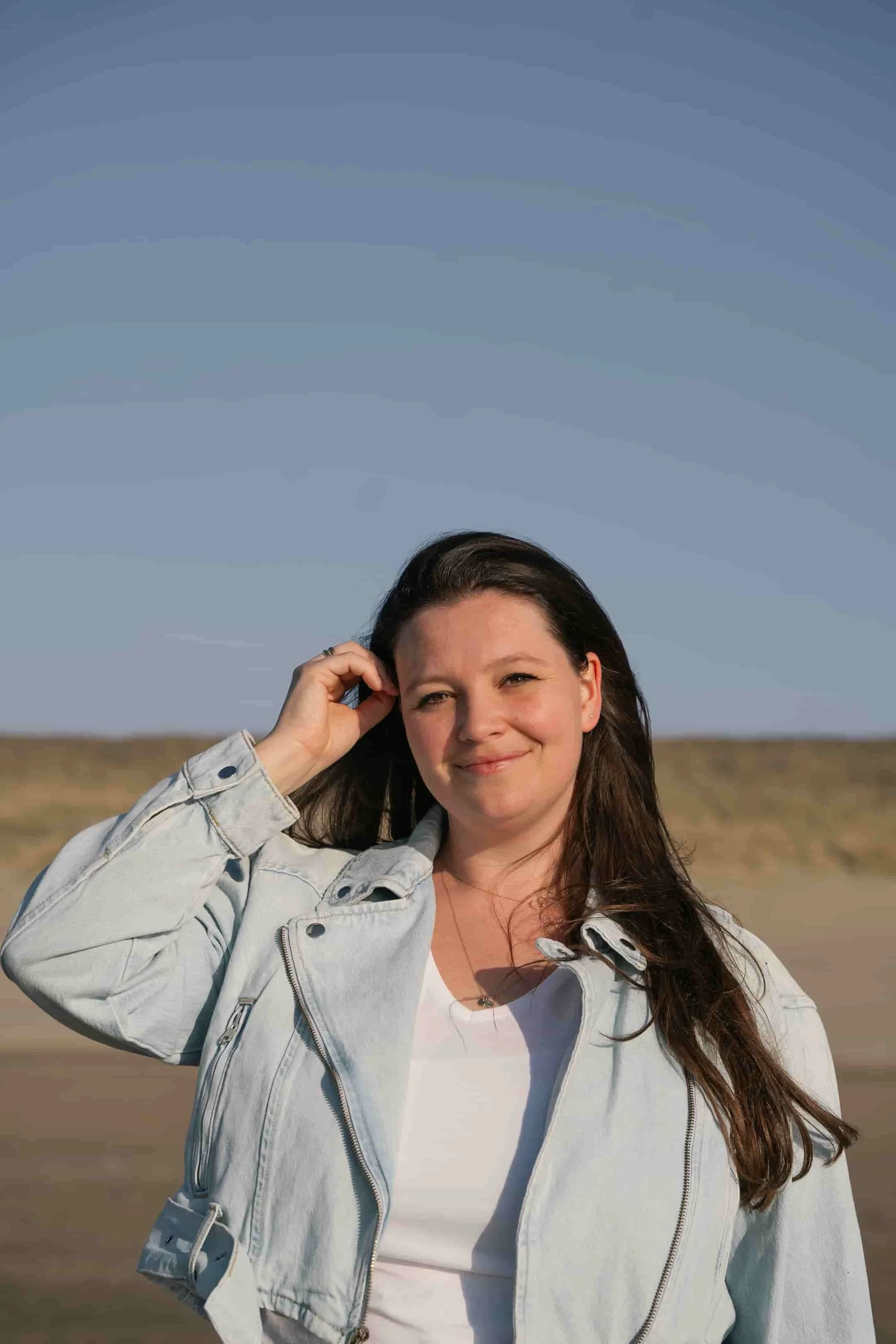 een jonge vrouw met lang, bruin haar die in een open veld staat, gekleed in een lichte spijkerjas en een witte top, onder een heldere blauwe lucht.