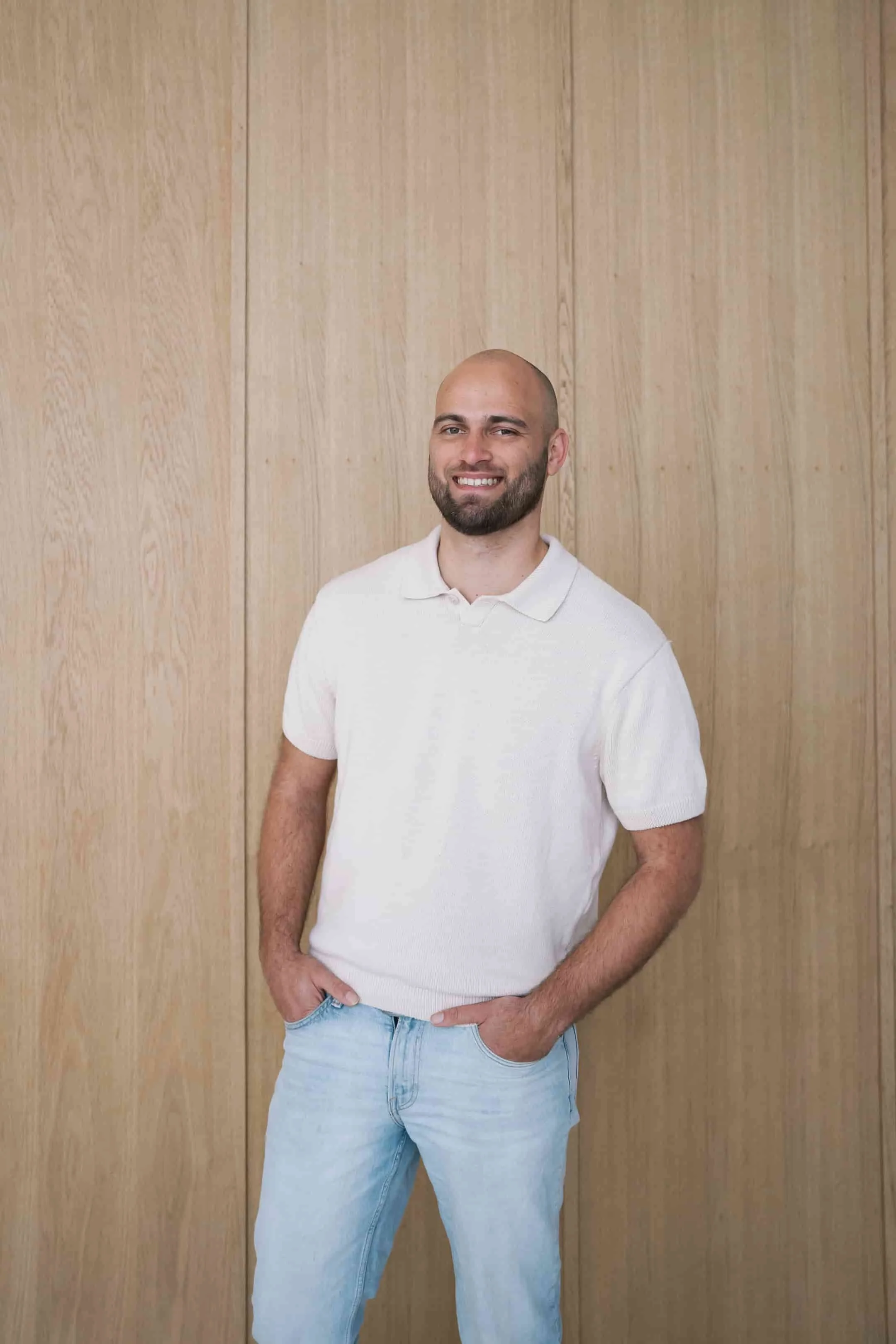 Een man met korte baard en een korte blond haarloze kop, draagt een witte polo en lichtblauwe jeans, staat voor een houten wand en glimlacht.