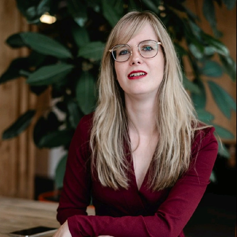 Een jonge vrouw met lang blond haar, brillen en rode lippen, zittend aan een tafel met een grote groene plant op de achtergrond.