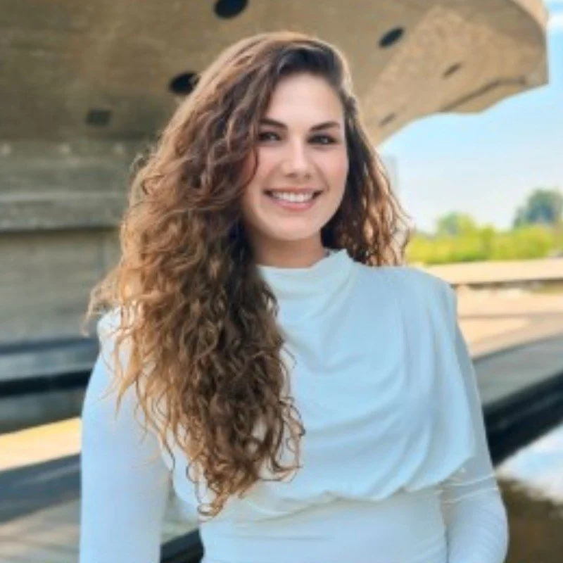 Close-up van een lachende vrouw met lang, krullend bruin haar, gekleed in een witte blouse, standing outdoors near water en een moderne brug.