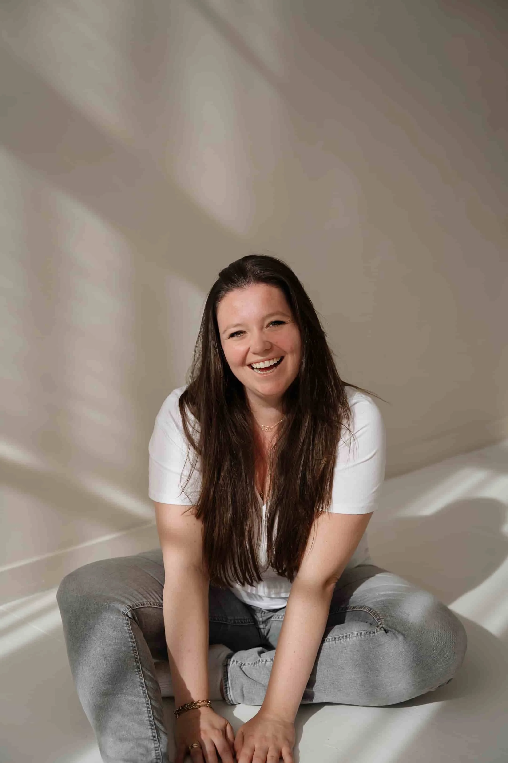 Een lachende vrouw met lang bruin haar zittend op de vloer, gekleed in een witte blouse en grijze spijkerbroek, voor een neutrale achtergrond met schaduwpatronen.