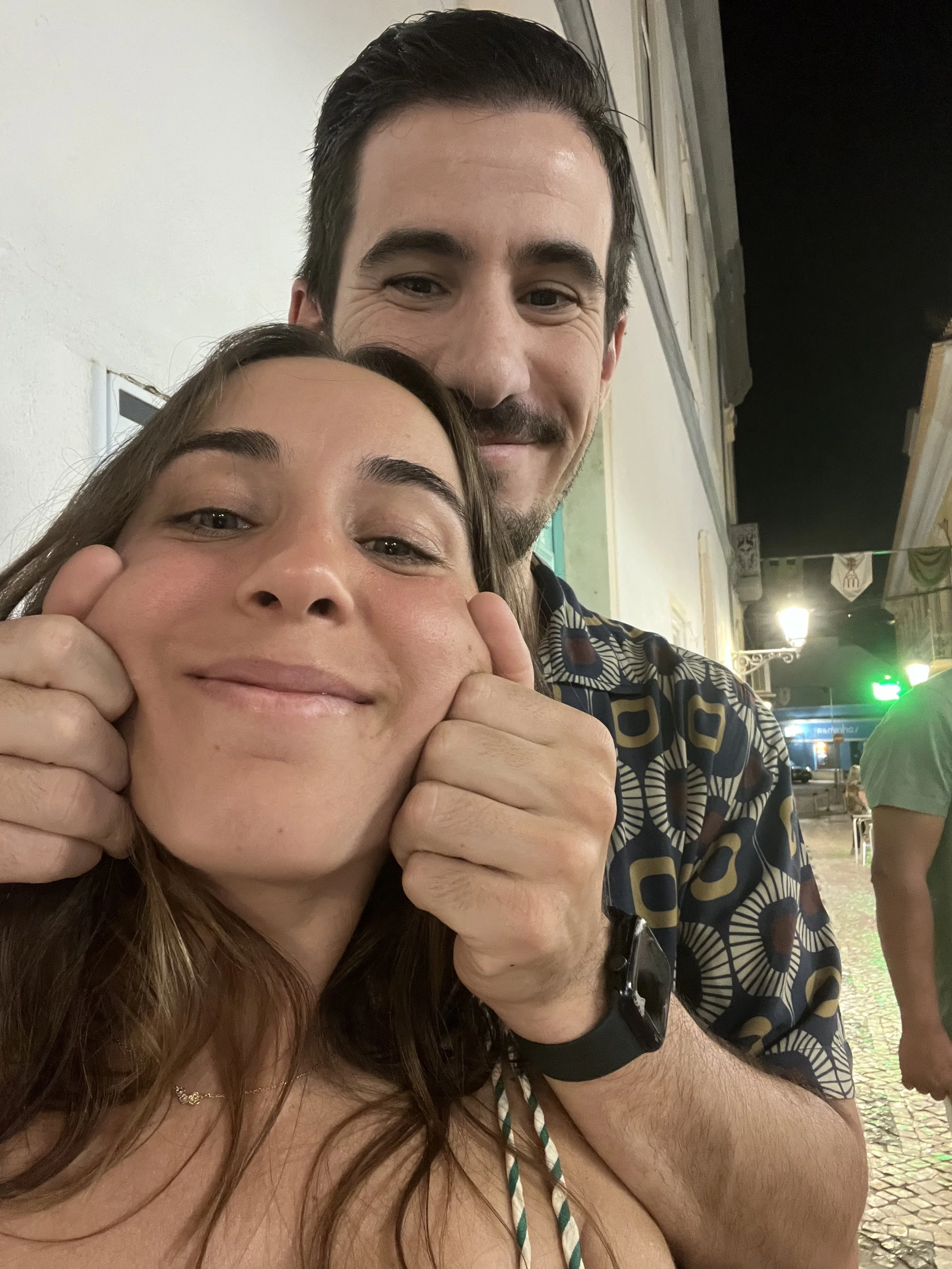 Um homem e uma mulher sorrindo para a câmera, em uma rua à noite, com o homem segurando a mulher pelos braços.