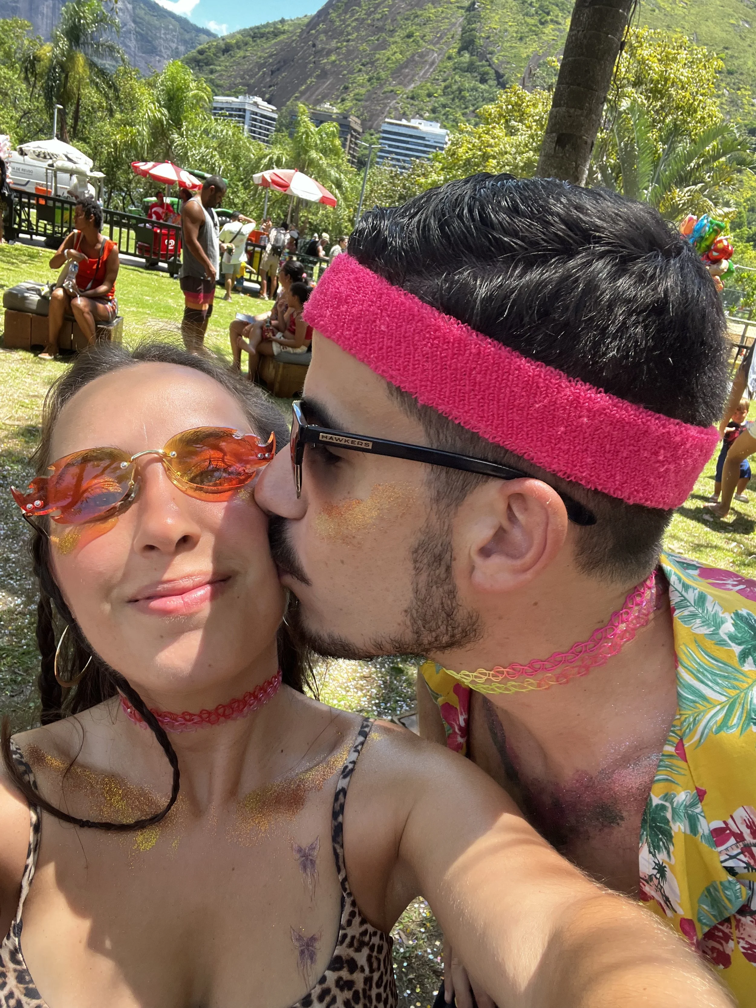 Casal se fazendo um beijo na bochecha em um parque com árvores e várias pessoas ao fundo, em um dia ensolarado.