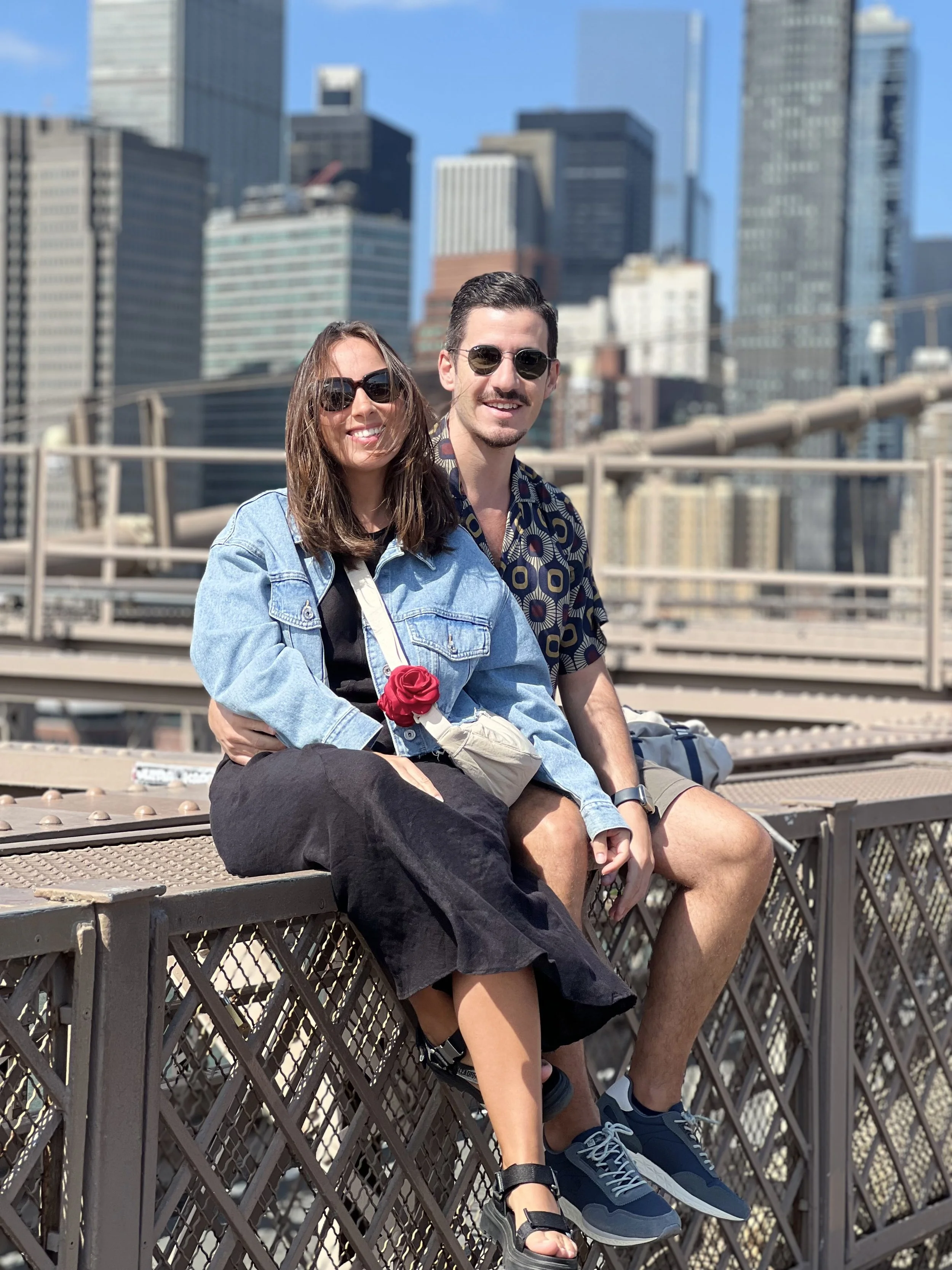 Um casal sentado na estrutura de uma ponte em uma cidade com arranha-céus ao fundo, ambos usando óculos de sol e sorrindo.